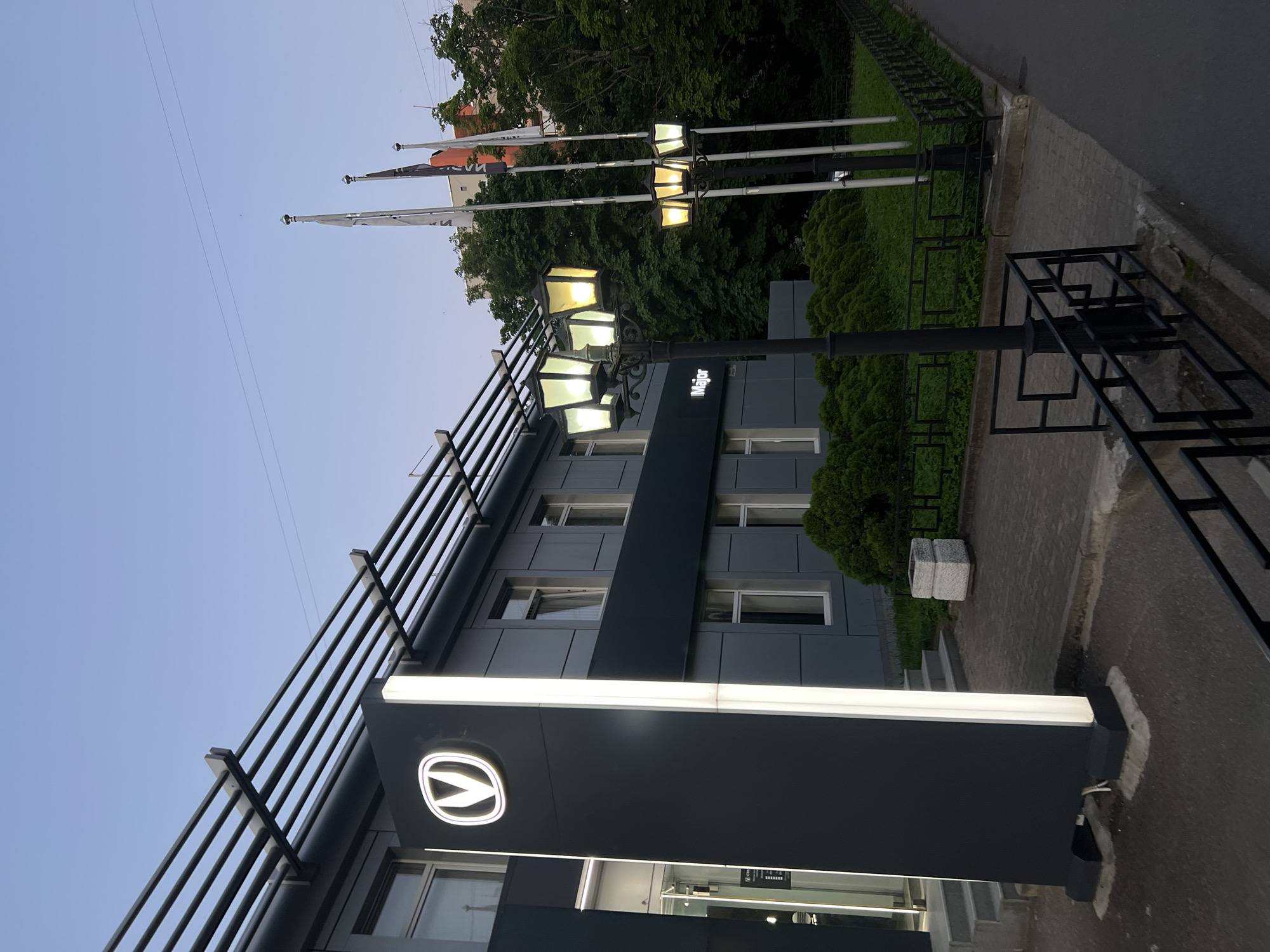 The image captures the exterior of a modern commercial building at dusk, located in Saint Petersburg, Russia. The two-story building features a dark gray paneled facade with white-framed rectangular windows. A prominent, tall, vertical pylon, illuminated along its edges, stands on the left side of the entrance, displaying a circular, illuminated "V" logo, which is the Changan Automobile emblem. Above the ground floor windows, the word "Major" is affixed to the building's facade, also illuminated. Extending horizontally from the roofline over the entrance is a series of parallel gray metal bars, forming a sunshade or decorative element.
In the foreground, two decorative, multi-lamp streetlights are active, casting yellow and white light. The streetlight closer to the building has five lamps, while the one further right has three. Adjacent to the streetlights are three white flagpoles, from which flags (partially obscured, but one appears black and another possibly red/orange) hang. The area in front of the building is landscaped with meticulously trimmed green hedges and a section of paved walkway, alongside an asphalt path or road on the right. A black metal fence or railing delineates the property line. In the background, lush green trees are visible, and a portion of another building with an orange/red facade can be seen. The sky is a clear, light blue, indicative of twilight, with a faint power line traversing the upper frame. No individuals are present in the scene.
The specific location is identified as the Major Auto Changan dealership at **Vyazovaya Ulitsa, 10, St Petersburg, Russia, 197110**.