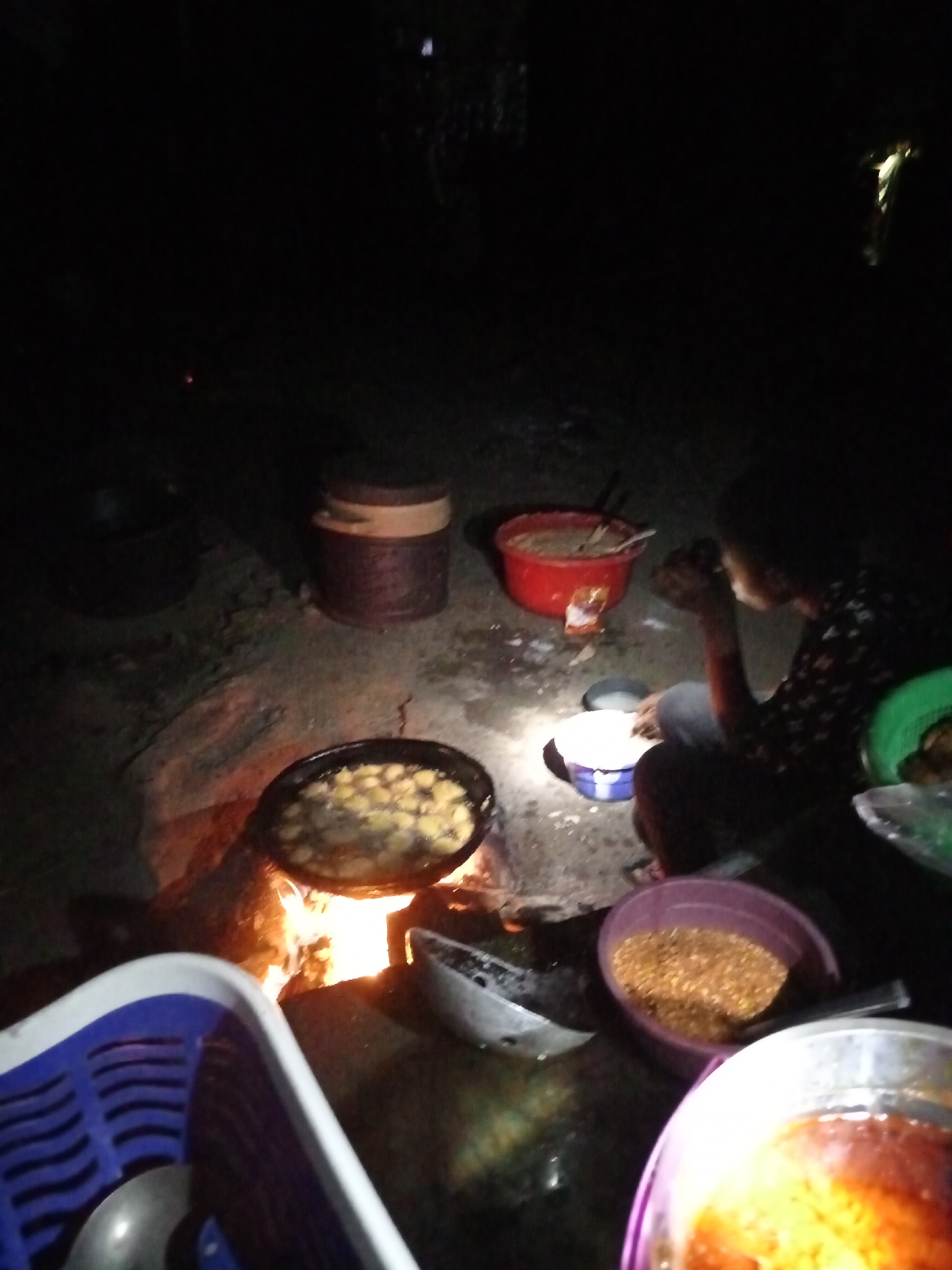 This media file captures an outdoor scene at night in Keffi, Nigeria, illuminated predominantly by an open cooking fire. The ground appears to be dirt or uneven terrain.
One person is visible on the right, seated on the ground with their side to the camera, their face obscured by shadow. They seem to be engaged in eating or preparing food.
The central activity revolves around cooking. A large black pot is placed directly over an open fire, with visible flames providing both heat for cooking and light for the scene. The pot contains light-colored food items, possibly fried dough or similar snacks, bubbling in liquid.
Several food containers are scattered around. A red bucket or bowl filled with a brownish substance and a spoon is located to the left of the seated person. A smaller white and blue bowl is positioned directly in front of the person, reflecting some light. Further right, a purple bowl holds a granular, yellowish-brown food, possibly roasted nuts or grains. In the foreground, partially visible, is another bowl containing a reddish liquid, likely a sauce or soup.
In the background, a large black pot and a tall, dark wooden barrel-like container are faintly discernible. A white and blue plastic basket with a ladle is partly visible in the lower left foreground.
The scene depicts a practical aspect of life, likely a communal meal preparation or gathering, with the fire serving as a crucial element for both sustenance and visibility in the darkness. There is no visible text in the image.