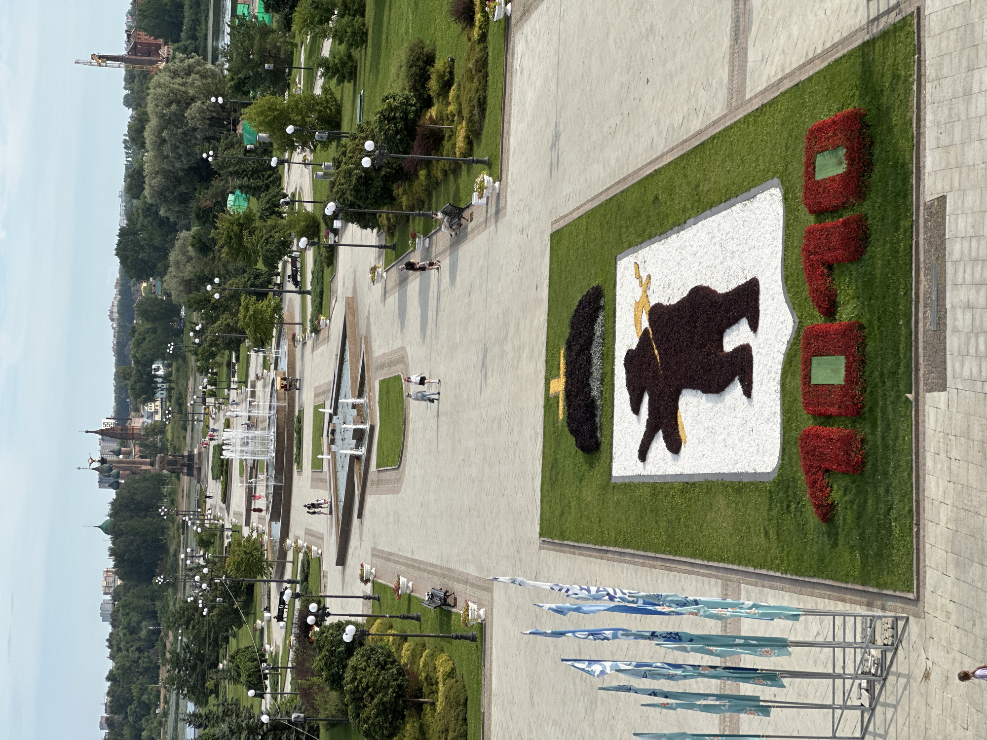 This is a high-angle, medium shot of a city park in Rusanovo, Russia, on a bright, slightly overcast day. The park features a large, paved walkway leading to a series of decorative fountains and manicured green spaces. In the foreground, a large flower bed depicts a bear silhouette with a yellow lightning bolt, standing on a white base, with the red floral numbers "2020" below. Several blue flags on poles are arranged to the left of the flower bed. The background reveals a cityscape with buildings, including churches with distinctive green and red domes, and a distant construction crane. People are seen walking and sitting on benches throughout the park, and a horse-drawn carriage is visible near one of the fountains. The environment is well-maintained, with lush trees and well-kept lawns.
