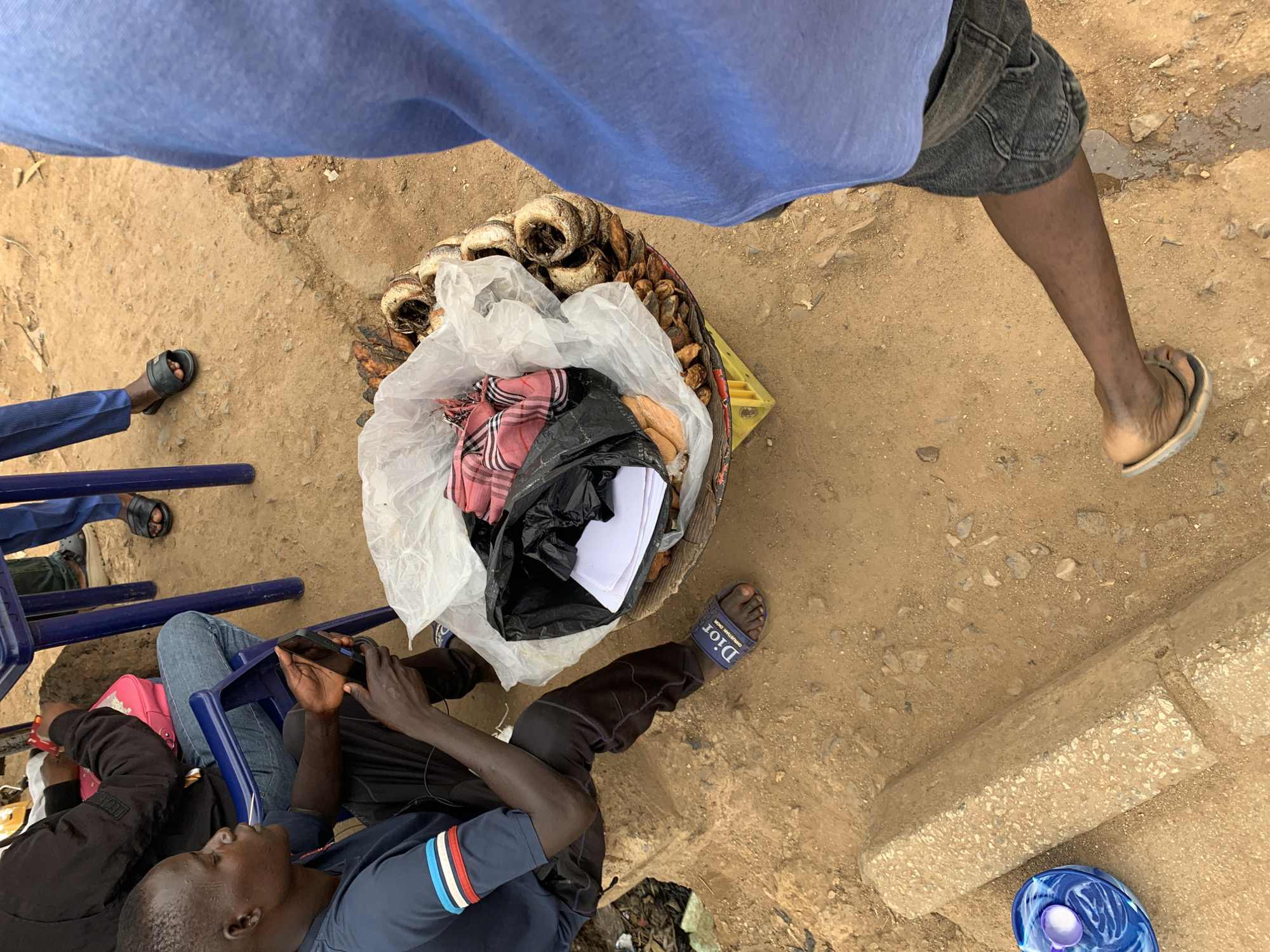 A high-angle shot shows a group of people in Rafin Par, Nigeria, sitting on blue plastic chairs with a basket of goods between them. The ground is dirt with some paving stones visible. A person in a blue shirt and grey shorts stands above the basket, partially obstructing the view. The basket contains dried fish, covered partially by white plastic and black bags. One person is holding a cell phone and looking at it, while another is looking away. A blue plastic bowl is on the ground near a curb. Some of the people are wearing sandals.