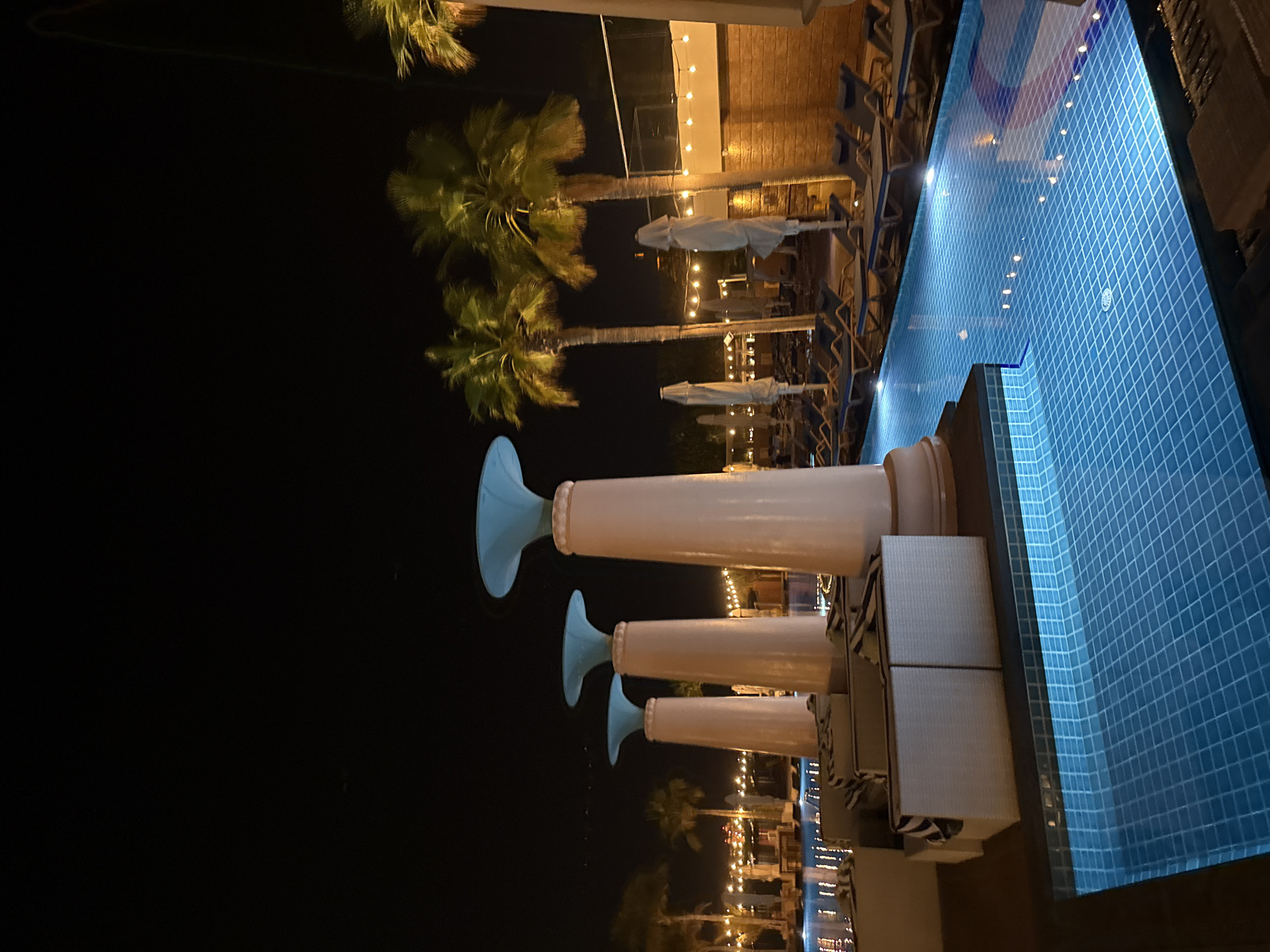 This is a nighttime photograph taken in Dubai, United Arab Emirates, depicting a resort-style swimming pool area. The pool itself is illuminated with blue lights, casting reflections on the tiled surface. Along the edge of the pool, there are rows of empty lounge chairs and closed white umbrellas. In the foreground, several white cushioned seating areas are arranged on a wooden deck. Further back, tall, light-colored pillars with blue conical tops line the pool area, spaced at intervals. Palm trees are visible behind the pillars, with their fronds illuminated by ambient lighting. In the background, a building with brickwork and string lights can be seen, suggesting a commercial or hospitality setting. The sky is dark and clear.