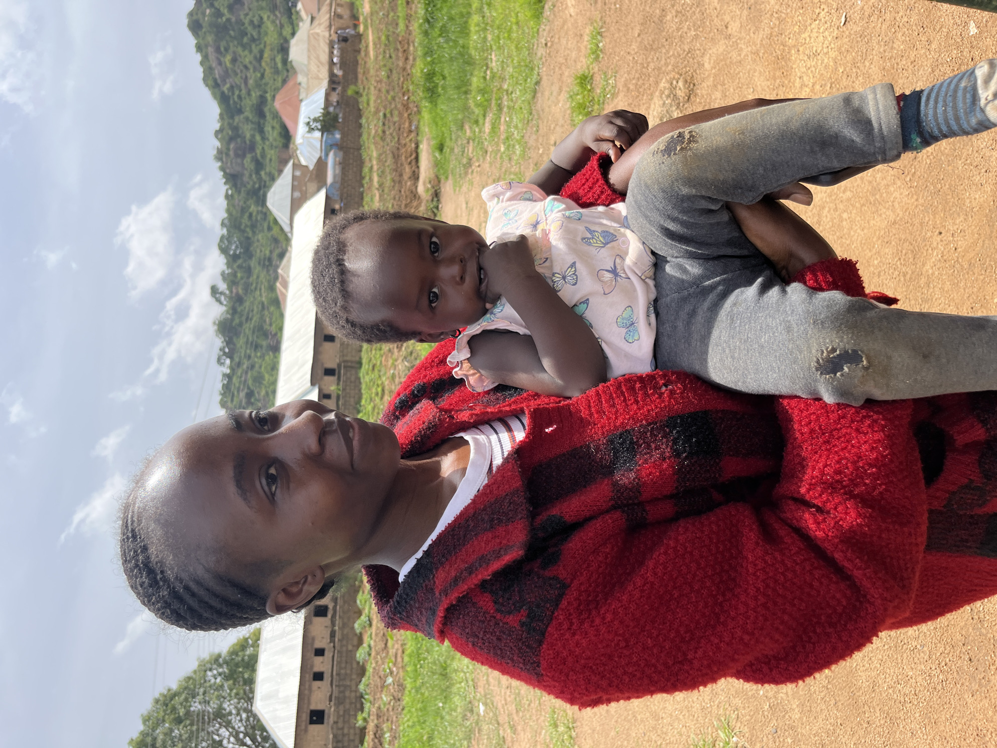 A young woman with braided hair holds a baby in her arms. She is wearing a red and black checkered sweater over a white and striped t-shirt. The baby is wearing a pink shirt with butterflies and grey pants. The background shows buildings and a grassy field, suggesting a rural or semi-rural setting, likely in Bukuru, Nigeria. The sky is overcast with scattered clouds.