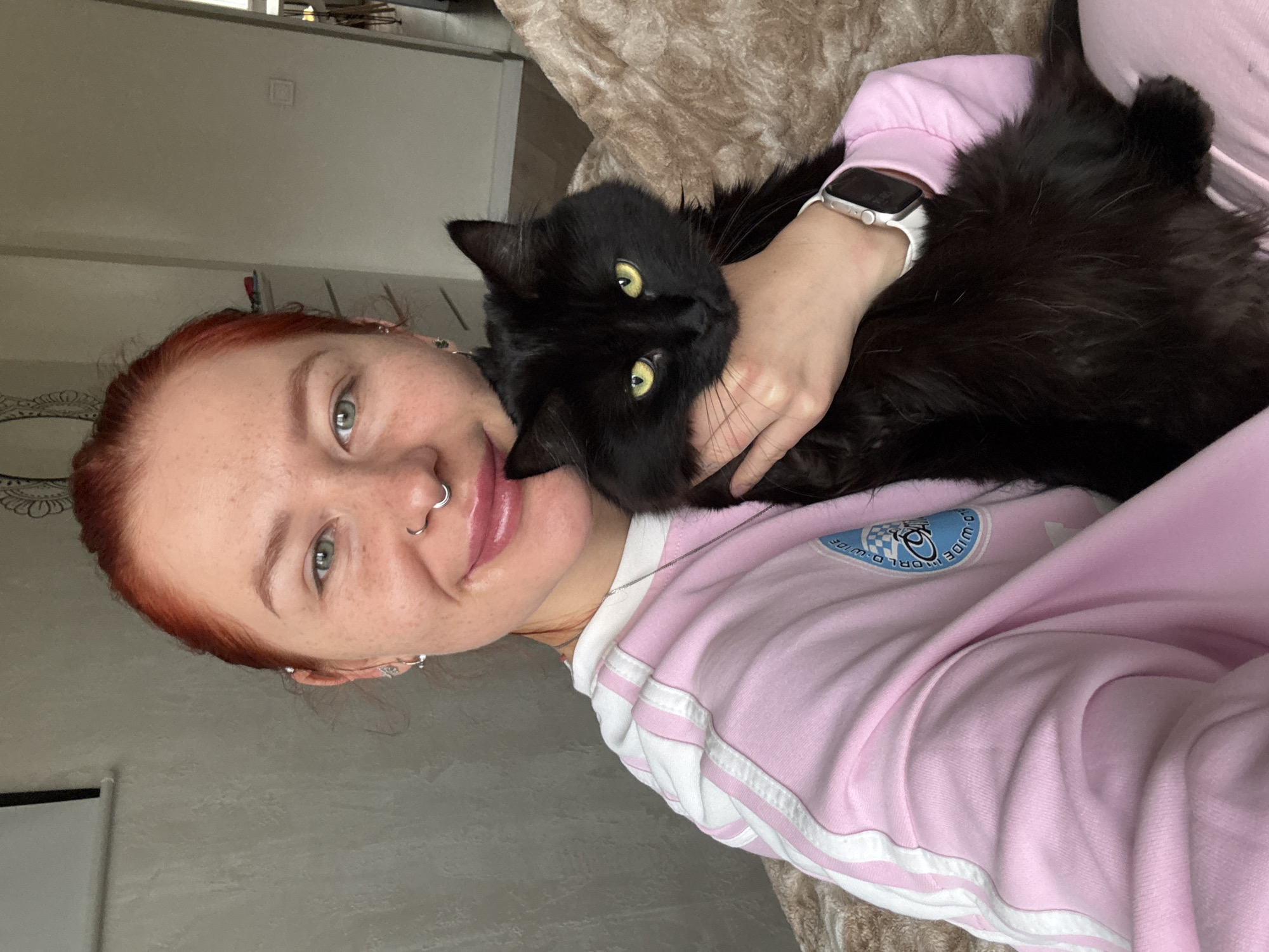 This vertical-format image, taken in Rostov-on-Don, Russia, depicts a woman and a black cat in a close-up, affectionate indoor scene. The woman, positioned on the left side of the frame, has reddish hair, green eyes, and light skin dotted with freckles. She offers a soft smile to the camera. She is wearing a light pink long-sleeved top that features white stripes on the sleeves and collar. A circular blue patch on her upper left chest displays a checkered pattern and partially visible text, "WORLD WIDE," characteristic of a Vans logo. A white smartwatch adorns her left wrist, and she has a nose piercing and earrings.
She holds a fluffy black cat with bright yellow-green eyes close to her, its head turned towards the viewer with a neutral yet engaged expression. The cat is comfortably nestled against her neck and chest, enveloped by her arm. The background reveals light-colored walls or ceiling and a textured brown blanket or throw, indicating a cozy home environment. The ambient lighting suggests it is daytime. The image conveys a sense of warmth, comfort, and the strong bond between the woman and her pet.