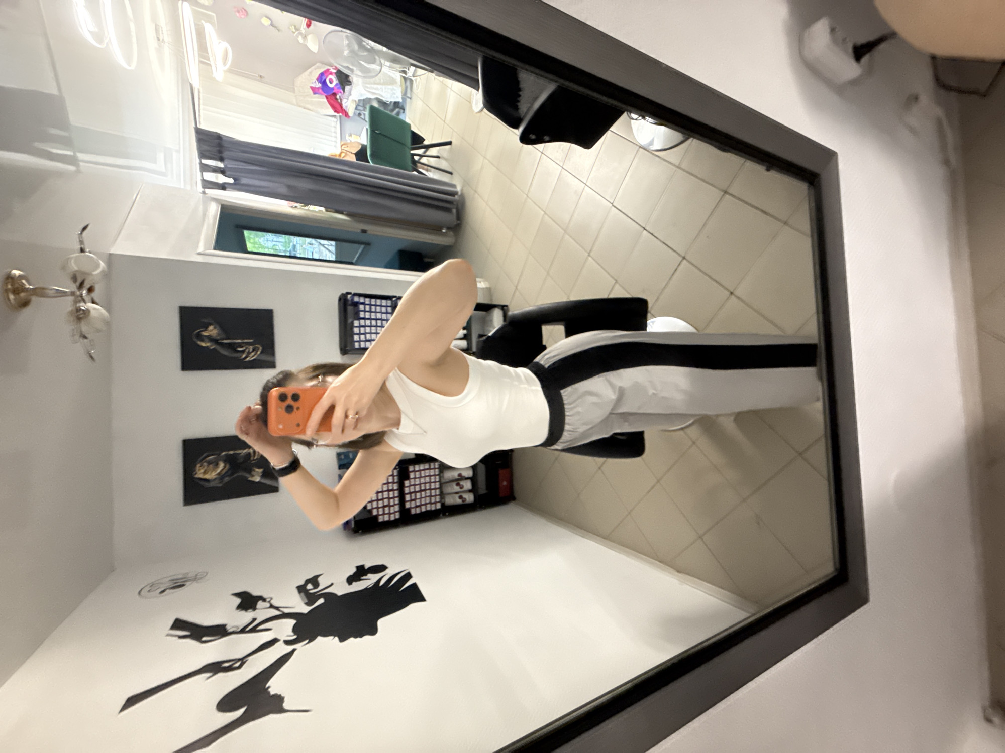 The image depicts a reflection in a framed mirror, likely taken by the person visible in the reflection. The reflection shows a woman holding an orange smartphone, presumably taking the picture. She is wearing a white tank top and grey sweatpants with black stripes on the sides. She is sitting in what appears to be a black salon chair.
The background of the reflection reveals aspects of the room: a white wall with black silhouette artwork, and some shelves with what might be salon products. The flooring has a light tile pattern. Through the mirror, we can see what may be another area of the salon, including salon chairs and a window with grey blinds.
Based on provided location metadata, this scene is in Chaikovsky, Russia, although nothing specific in the image confirms this.