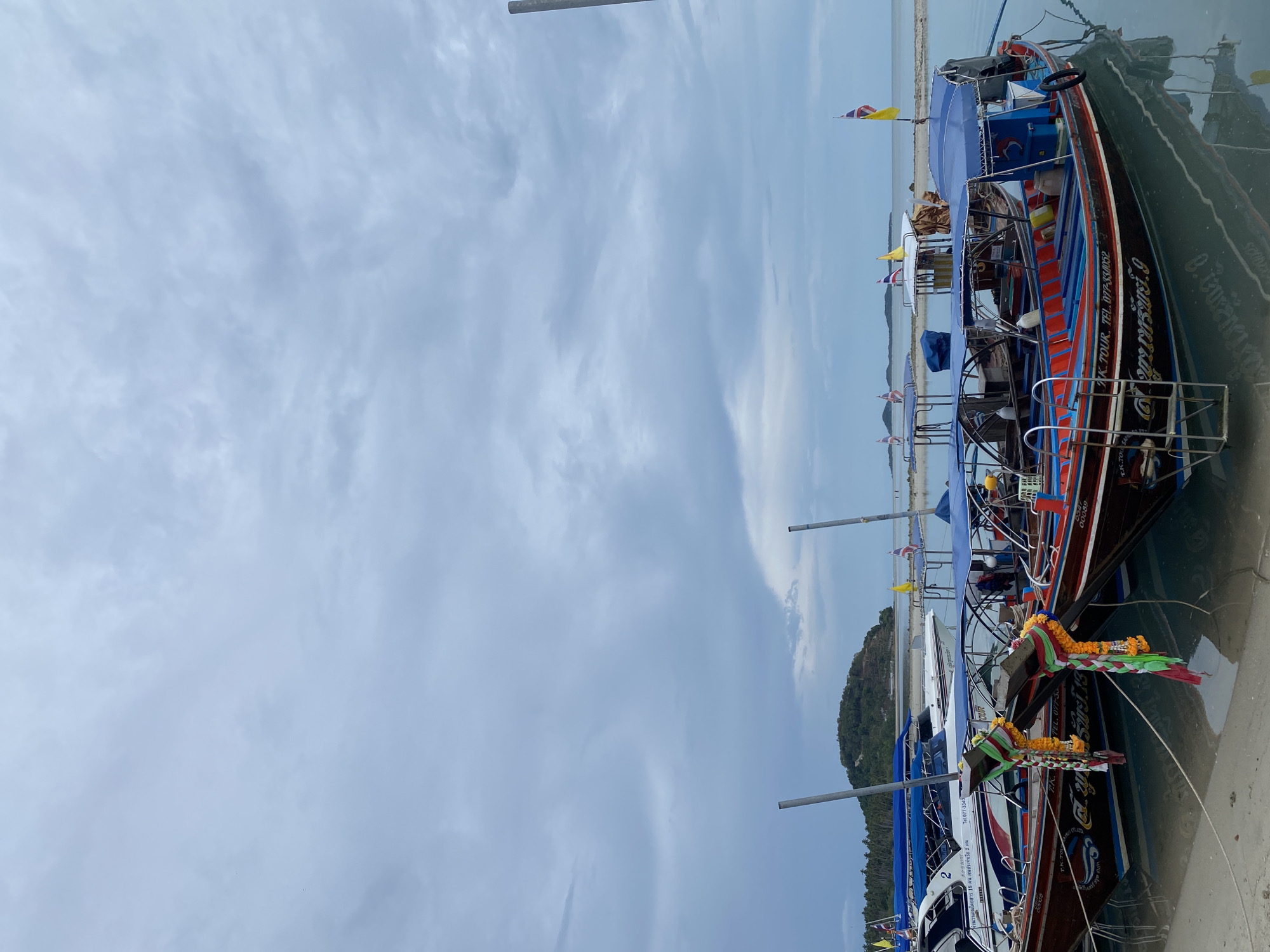 The image shows several Thai longtail boats docked at a shore, likely in Baan Thong Krut, Thailand. The boats are vividly decorated, with blue canopies and colorful flags. Some boats have garlands hanging on the bows. The sky is overcast with light clouds. The boats appear to be used for tourism purposes, based on the markings and decorations.
