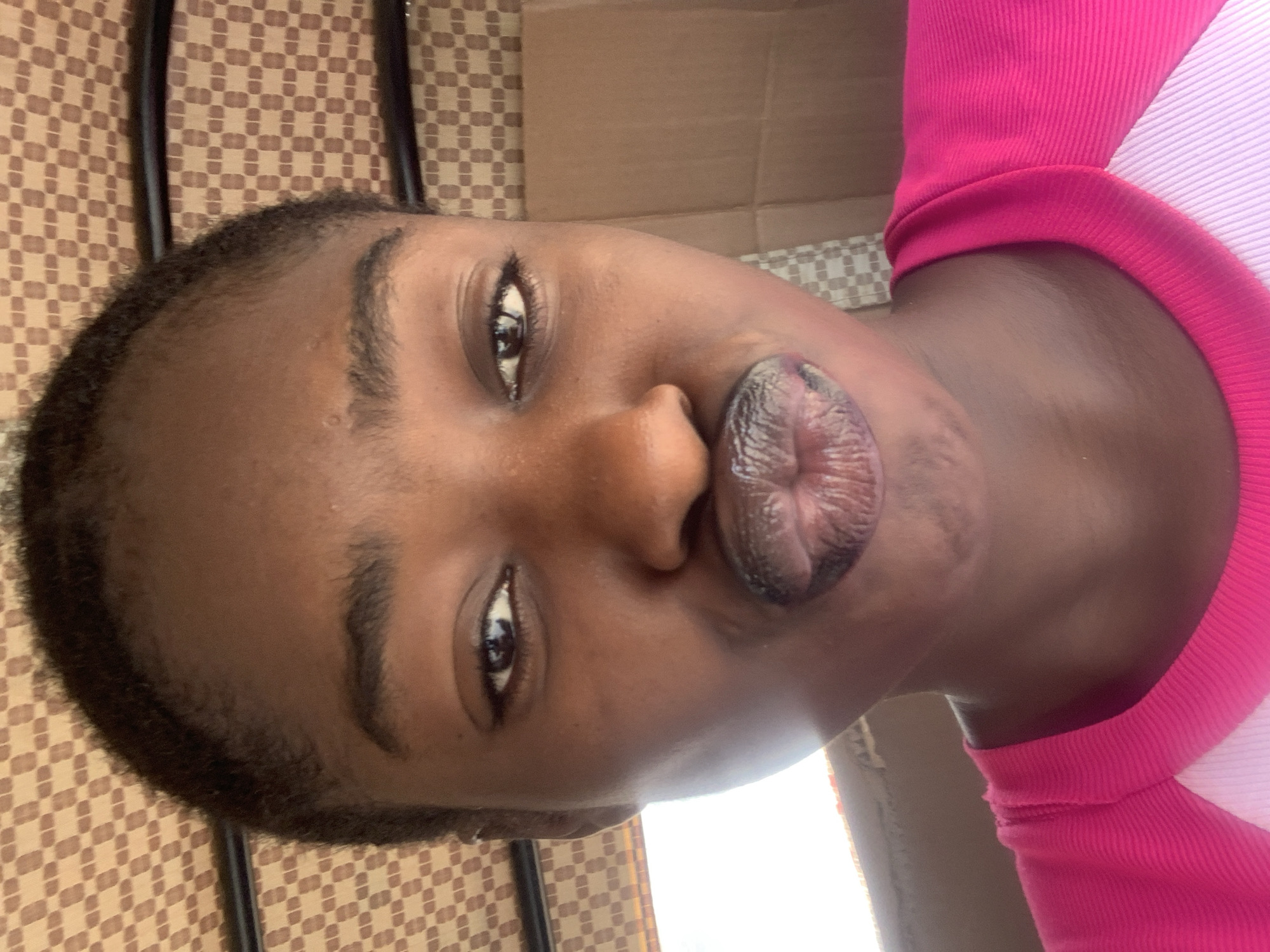 A close-up, rotated view of a person with dark skin, short dark hair, and brown eyes. The person is puckering their lips. They are wearing a pink and white shirt. The background shows a brown checkered surface and a piece of cardboard. The location of Rafin Par, Nigeria is not discernible from the image.