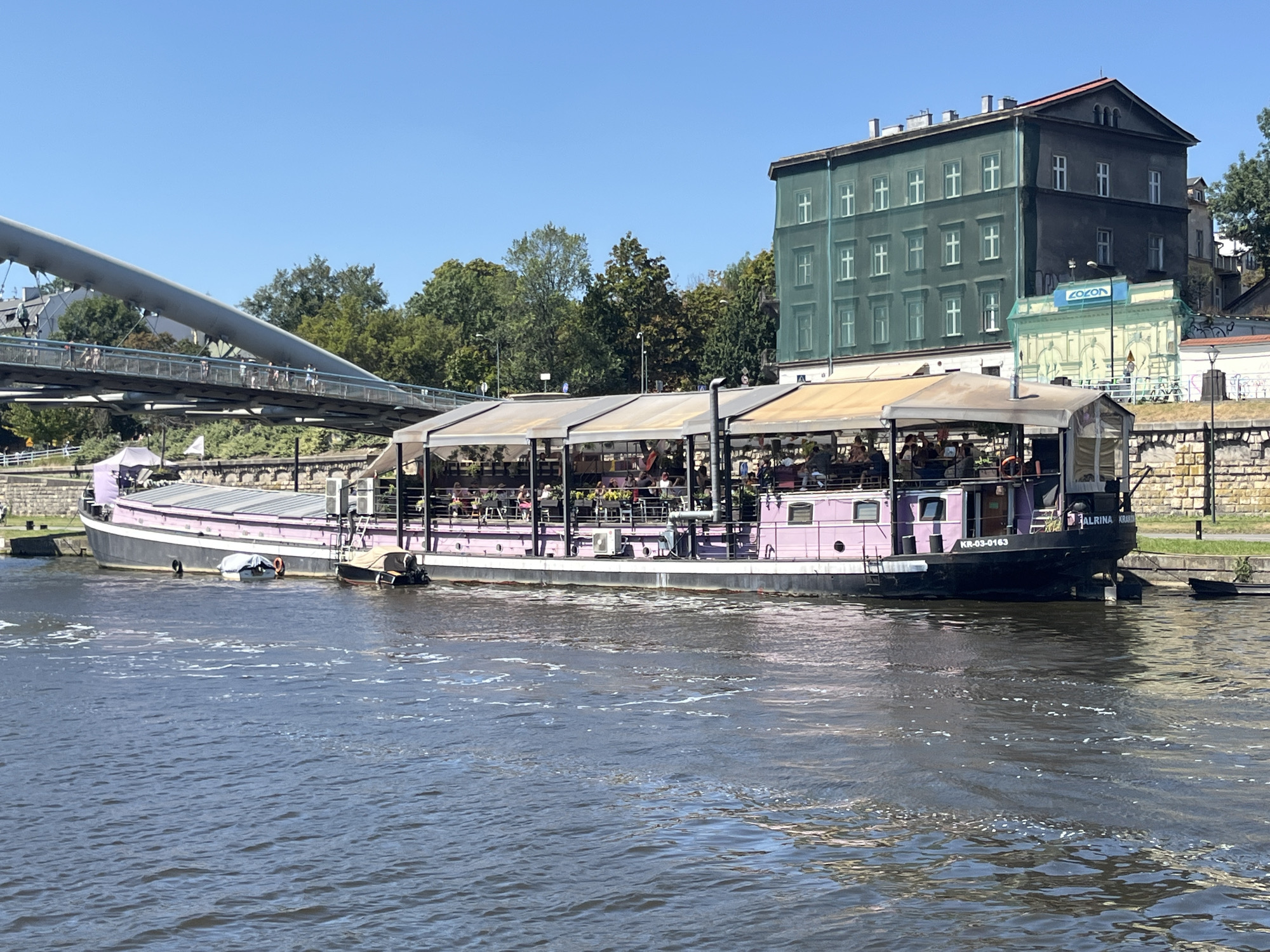 In Krakow, Poland, a flat-bottomed barge with a pink hull and a dark superstructure is moored on the Vistula River. The barge is operating as an open-air restaurant, with tables and patrons visible under a white canopy. The boat bears the inscription "ALBINA KRAK." and the registration number "KR-03-0163". Behind the barge, a modern pedestrian bridge with people walking across it spans the river. Further in the background, buildings are visible, including a multi-story structure covered in green netting, suggesting renovation or construction. The sky is clear and blue, and the water is calm with some ripples.