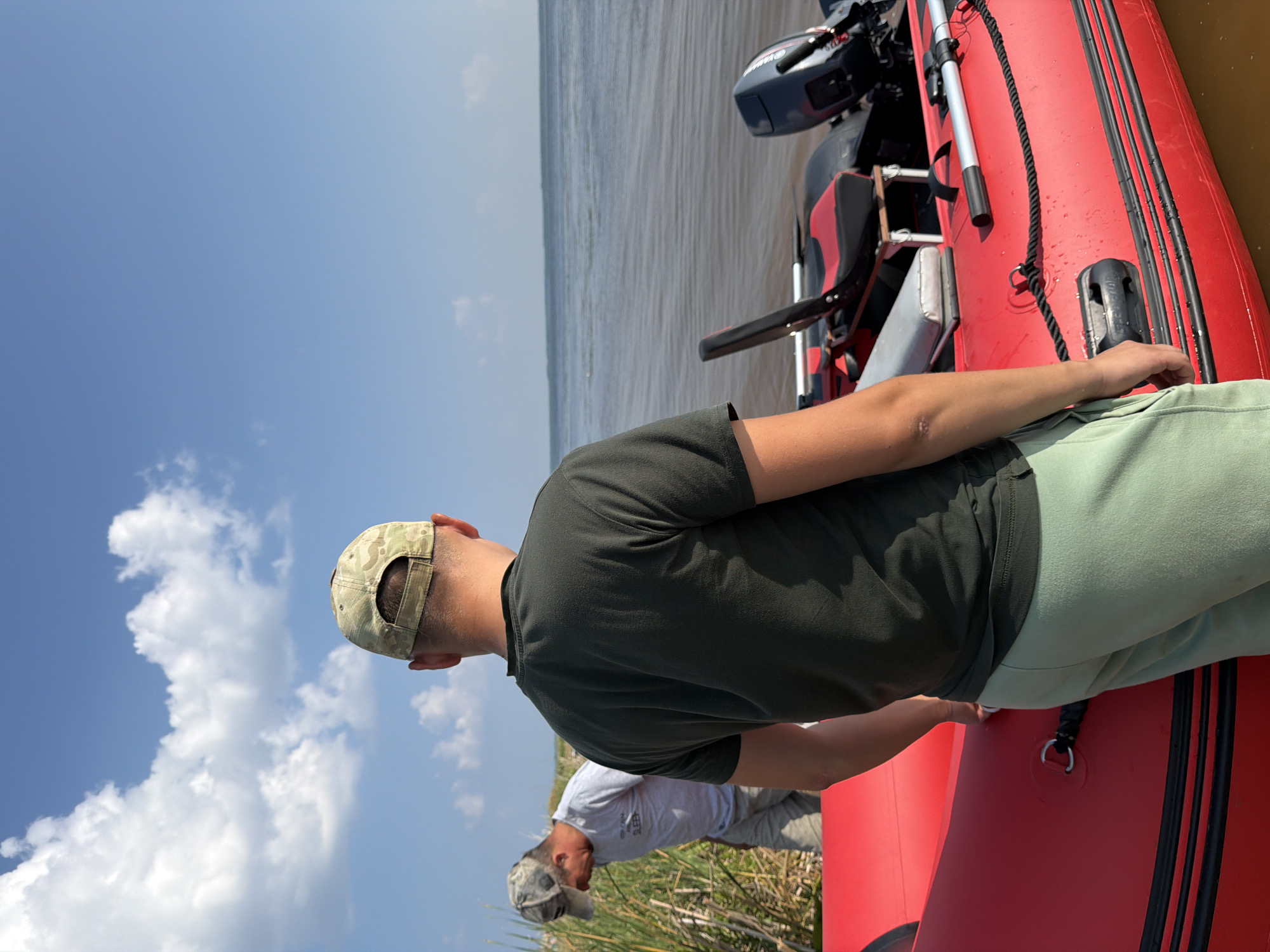 Two adult males are on the shore of a body of water, possibly a river or lake, with a red inflatable boat equipped with a motor. The man in the foreground, wearing a dark green t-shirt and light green sweatpants, faces away from the camera, looking towards the water and the boat. He is wearing a camouflage baseball cap. The second man, further in the background and to the left, is also wearing a camouflage baseball cap and a light gray t-shirt with text on it. He is looking down towards the water and reeds. The sky is blue with scattered white clouds. The environment appears to be a natural, undeveloped area. The image was taken in or around Krasny Bor, Russia, based on the user's request.