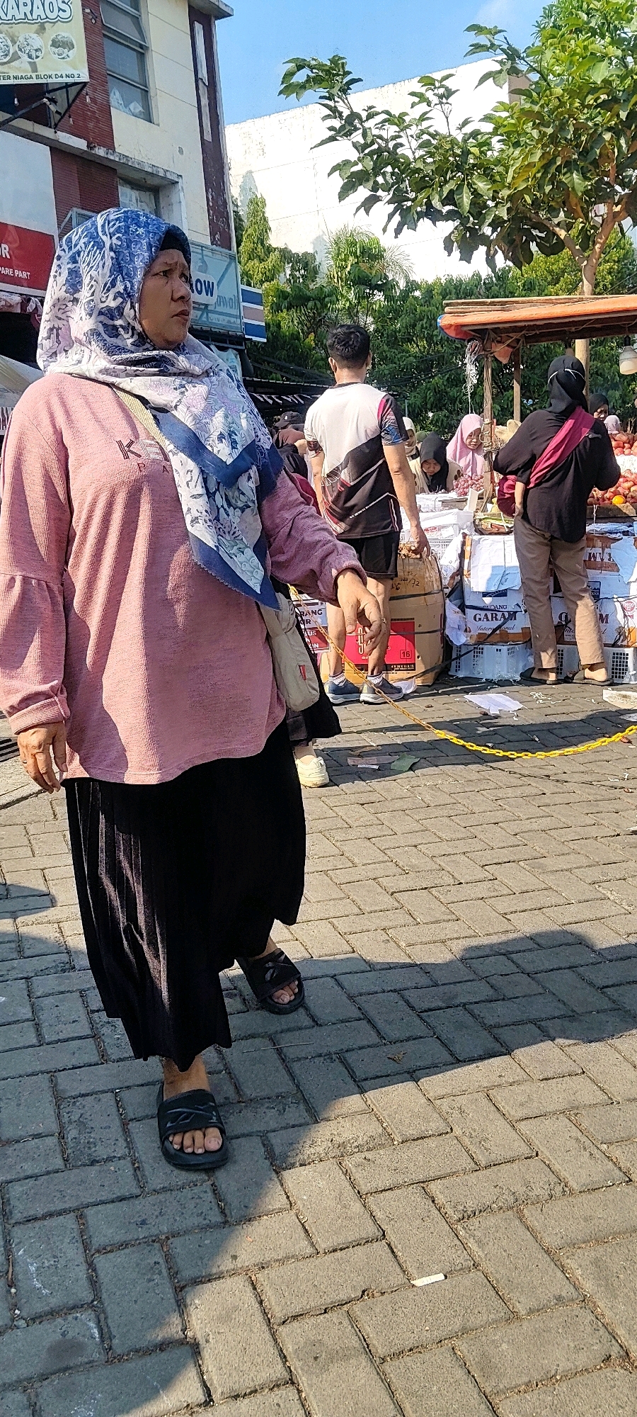 In the city of Cibatu, Indonesia, a street market scene unfolds. The foreground is dominated by a woman wearing a pink long-sleeved shirt and a black pleated skirt, with a blue and white patterned hijab. She is walking to the left, holding a cigarette in her right hand. Behind her, a man in a white and black t-shirt and black shorts stands facing away from the camera, carrying a large cardboard box. Further back, other individuals are visible among stalls laden with goods. One stall displays what appears to be fruit, with a woman in a black hijab and pink sash standing beside it. The ground is paved with interlocking gray bricks, casting shadows from the midday sun. The background features buildings, trees, and a clear blue sky. A yellow rope, possibly marking a boundary, runs across the paved area in front of the market stalls.