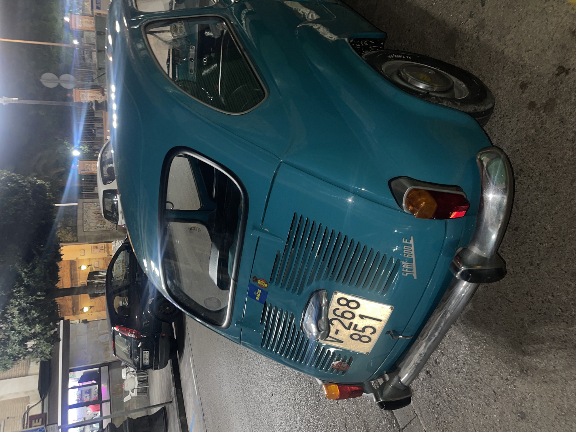 The image depicts a nighttime street scene in Manises, Spain. A teal blue-green SEAT 600 E classic car is prominently featured in the foreground, parked at an angle, viewed from its rear-left side. The car displays a Spanish license plate, "V-268-851," illuminated, and "SEAT 600 E" embossed on its lower rear panel, above a chrome bumper. A "RACE" sticker and a shield emblem are visible above the license plate. The car's interior is partially discernible through the rear-left window, showing the dashboard and steering wheel.
In the mid-ground, a dark-colored modern hatchback car, with the license plate "5198 FJB," is parked further down the street. Behind it, a commercial building with large windows is illuminated, revealing a person in a red shirt inside. White outdoor tables and chairs are arranged on the sidewalk in front of this building. A mature tree with dark foliage stands between the cars and the building.
Further in the background, to the right, a white car is partially visible. Beyond that, a decorative low wall or fence with patterned tiles and ornate pillars lines what appears to be a public space or park. The scene is lit by multiple artificial light sources, creating reflections on car surfaces and highlighting elements of the street and buildings. The ground consists of asphalt road and concrete sidewalk. No direct human interaction or explicit actions are depicted; the scene is static.