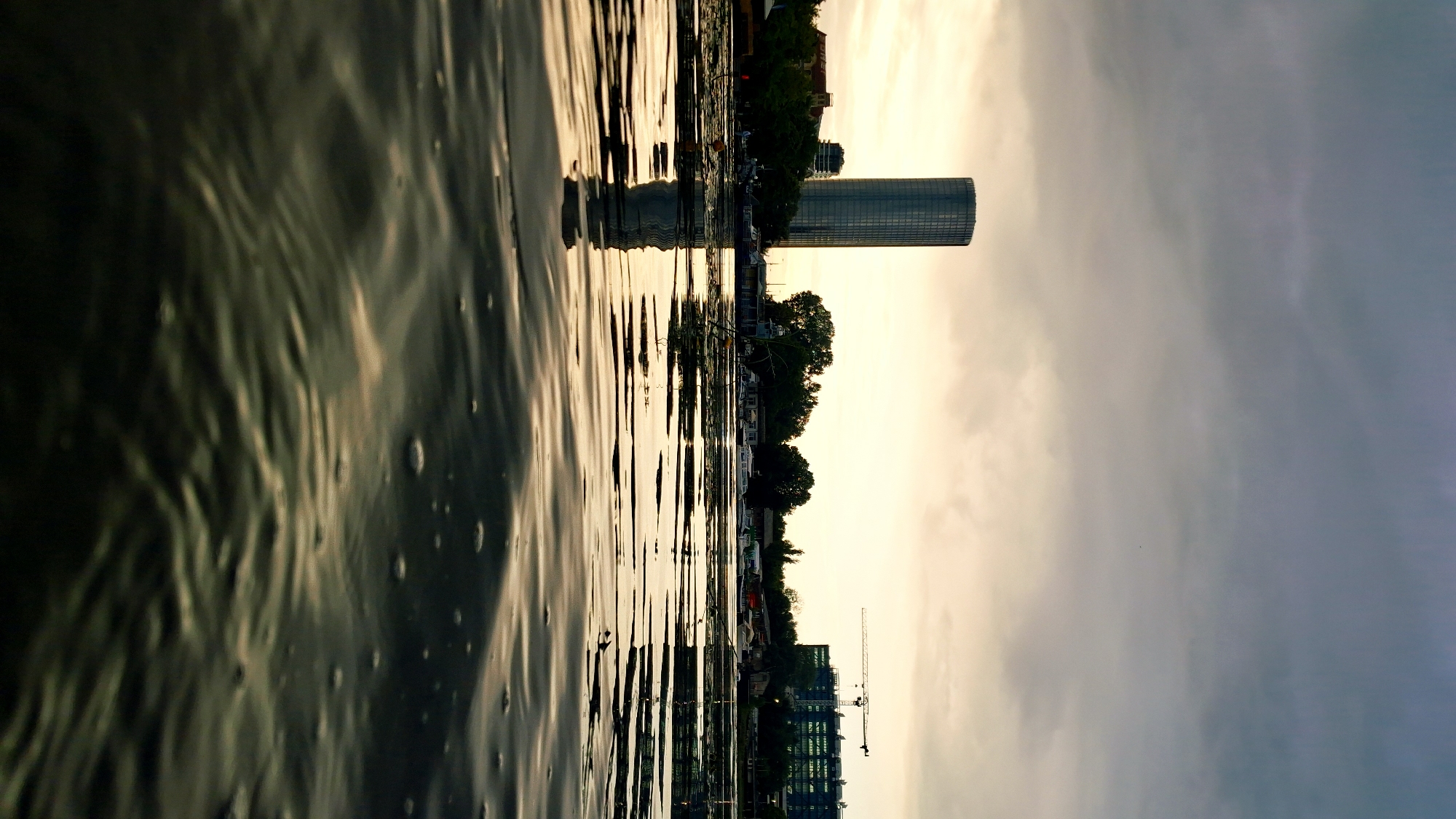 The image captures a low-angle view of the Daugava River in Riga, Latvia, during what appears to be sunset or overcast twilight. The foreground is dominated by the rippling surface of the river, with reflections of the sky and distant objects. The mid-ground reveals the cityscape, characterized by a prominent cylindrical skyscraper, likely the Z Towers, on the left. To the right, a large building complex is visible, with a construction crane positioned above it, indicating ongoing development. The shoreline is lined with trees and boats docked along the river. The sky is filled with dramatic, heavy clouds, with a subtle gradient of light suggesting the sun is low on the horizon. There are no discernible people or specific actions taking place. The overall atmosphere is muted and serene, with a focus on the interplay of water, architecture, and sky.