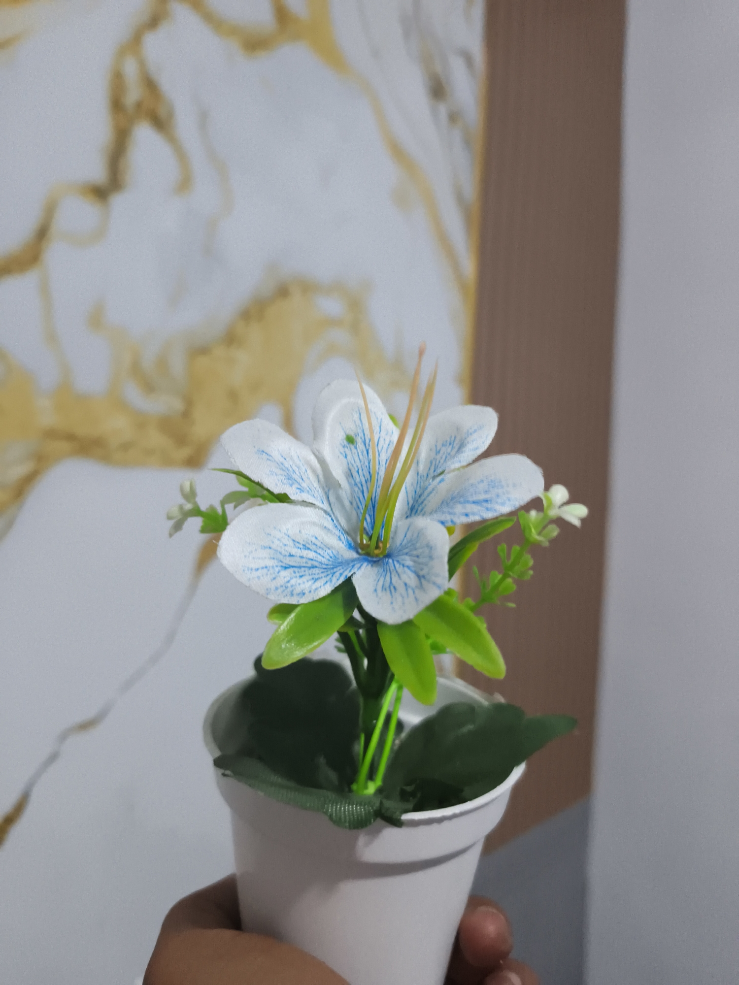 A close-up, eye-level shot shows a person's hand holding a small, white, potted artificial flower with white and blue petals and green leaves. The flower is positioned in the foreground, with a blurred background featuring a marbled wall with gold veins and a plain brown wall. The setting appears to be indoors, likely in a home or office environment in Pangkalpinang, Indonesia, given the context of the image request, though this cannot be definitively confirmed from visual cues alone.