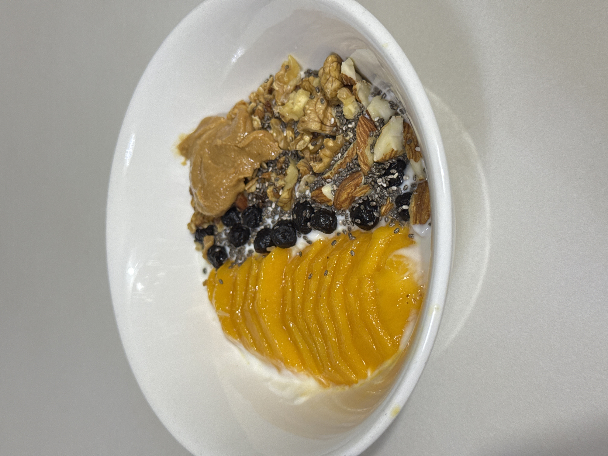 A white ceramic bowl contains a healthy breakfast. The bowl is filled with yogurt, topped with sliced mangoes, blueberries, chia seeds, walnuts, and almonds. A dollop of peanut butter sits on top of the nuts. The bowl is placed on a white, textured surface. The lighting is soft and diffused, suggesting an indoor setting. The background is a plain, light gray wall. There are no people visible in the image. The location is not specified in the image, and therefore cannot be determined to be Davao City, Philippines.