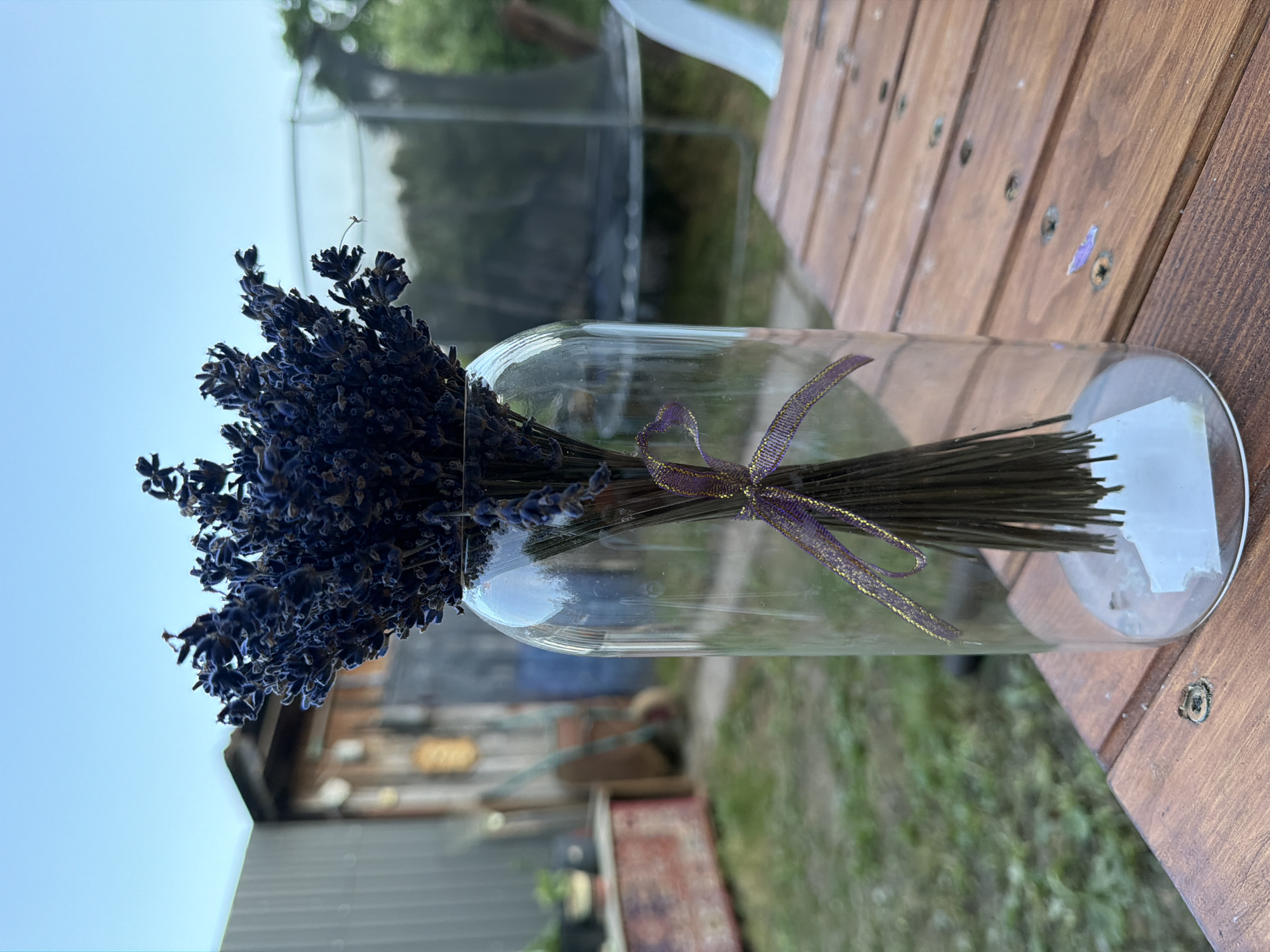 A bunch of dried lavender is displayed in a clear glass vase, tied with a purple and gold ribbon. The vase sits on a wooden outdoor table. In the background, there is a grassy yard with a trampoline, a fence, and some sheds. The sky is clear and blue. The location is the city of Kamianka-Buzka, Ukraine. There are no people or animals visible in the image.
