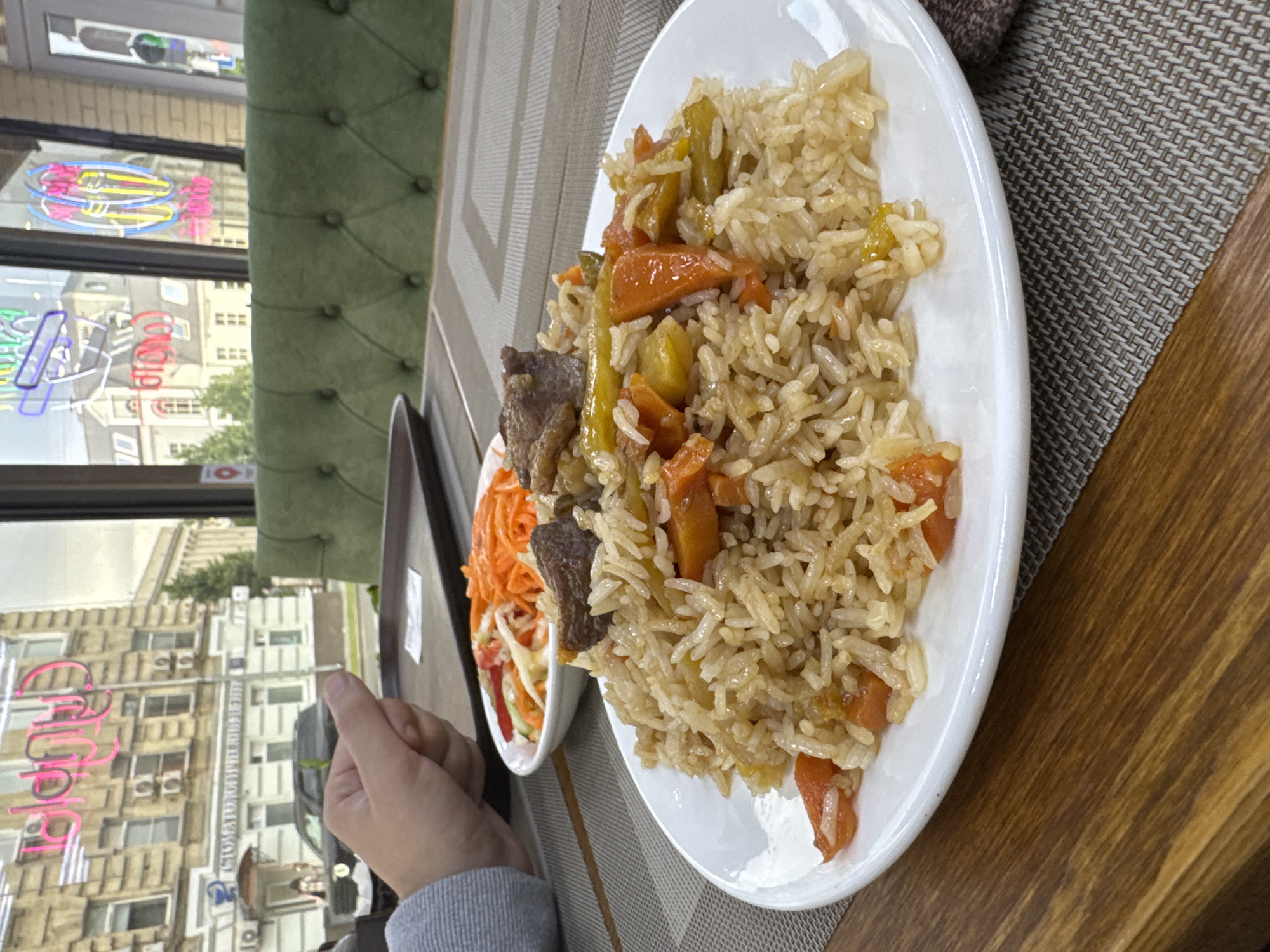 A close-up, eye-level shot shows a plate of pilaf with carrots and meat, accompanied by a side salad of shredded carrots, cucumbers, and red cabbage, on a table in a restaurant. The pilaf, a staple dish in Central Asian cuisine, is served in a white ceramic plate and is the central focus of the image. The salad is in a smaller white bowl and sits behind the pilaf. A hand, with the thumb slightly raised, is visible on the left side of the frame, suggesting a person is about to eat or has just finished eating. The table is covered with a woven placemat. In the background, a green tufted sofa is visible, and through a window, a city street scene in Moscow can be observed, with buildings, neon signs, and a parked car. The overall impression is of a casual dining experience in an urban setting.