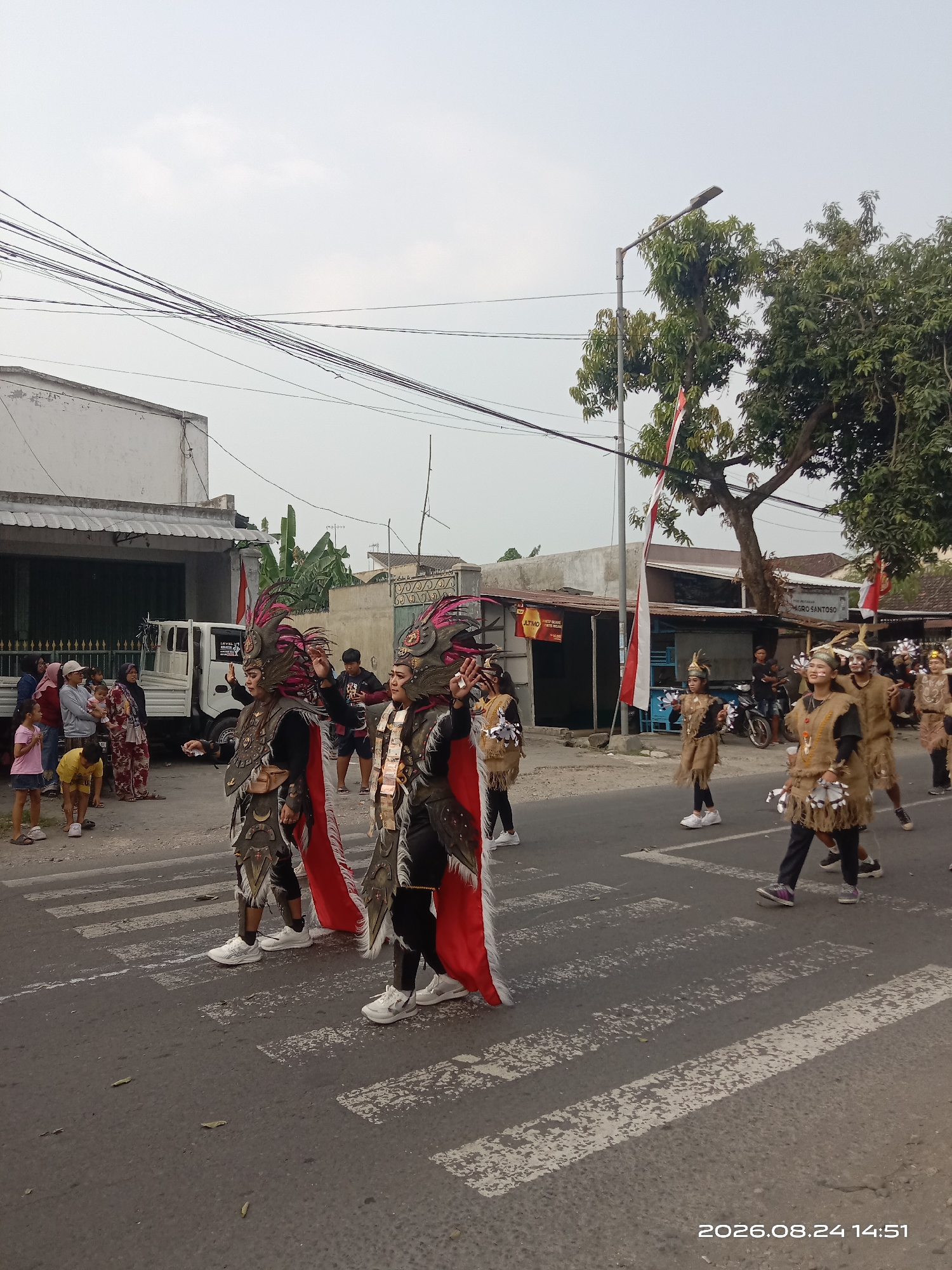The image captures a street scene in Gudo, Indonesia, likely during a cultural parade or festival. In the foreground, two individuals in elaborate, warrior-like costumes walk across a pedestrian crossing. Their costumes feature ornate headpieces with feathers, metallic chest armor, and flowing red capes. They are positioned on the right side of the frame, facing the viewer's left. To their right, other participants in similar, though less elaborate, costumes are visible further down the street, walking in the same direction.
On the left side of the street, a group of spectators are gathered, watching the procession. They consist of men, women, and children of various ages. A white truck is parked behind the spectators. The street is paved, and the pedestrian crossing is marked with white stripes. Buildings line the left side of the street, appearing to be shops or houses. Trees and streetlights are present along the right side of the street, with Indonesian flags displayed on poles. The sky is overcast, suggesting a cloudy day. The timestamp "2026.08.24 14:51" is visible in the bottom right corner of the image.