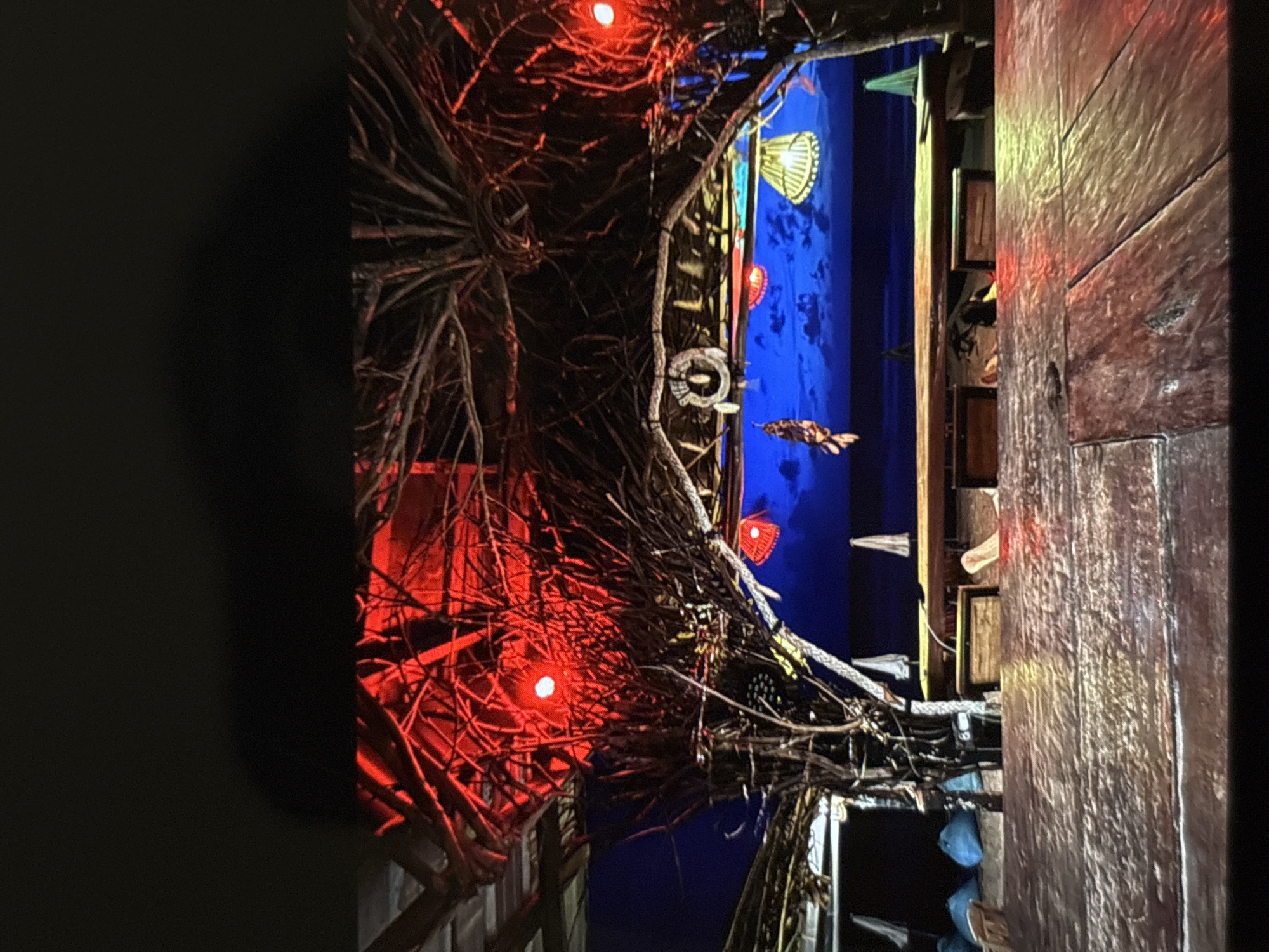 The image depicts a dimly lit outdoor scene, likely a bar or restaurant, with a focus on rustic decor and ambiance. The foreground is dominated by a weathered wooden table, its surface reflecting ambient light. Behind the table, a structure with a thatched roof is visible, supported by branches and adorned with red lights and hanging decorations, including what appears to be a dreamcatcher. The background reveals a deep blue, possibly nighttime, sky with clouds, and further back, silhouettes of beach umbrellas and tables. The overall impression is one of a tropical, possibly beachside, establishment in Chalong, Thailand, characterized by its natural materials and atmospheric lighting. There are no discernible people in the image.