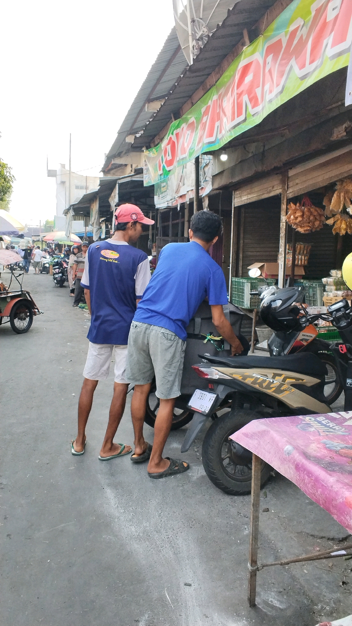 Two men stand on a paved road in front of a market stall in Mojokerto, Indonesia. The man on the left wears a blue and grey shirt, white shorts, and a red baseball cap. He faces away from the camera, looking towards the right. The man on the right wears a blue t-shirt and grey shorts. He is also facing away from the camera, leaning towards a motorcycle behind him. The motorcycle is black and gold. Behind the men, the market stall has a green banner with red text, and there are items hanging from the stall. Several other motorcycles and people are visible in the background. The sky is overcast.