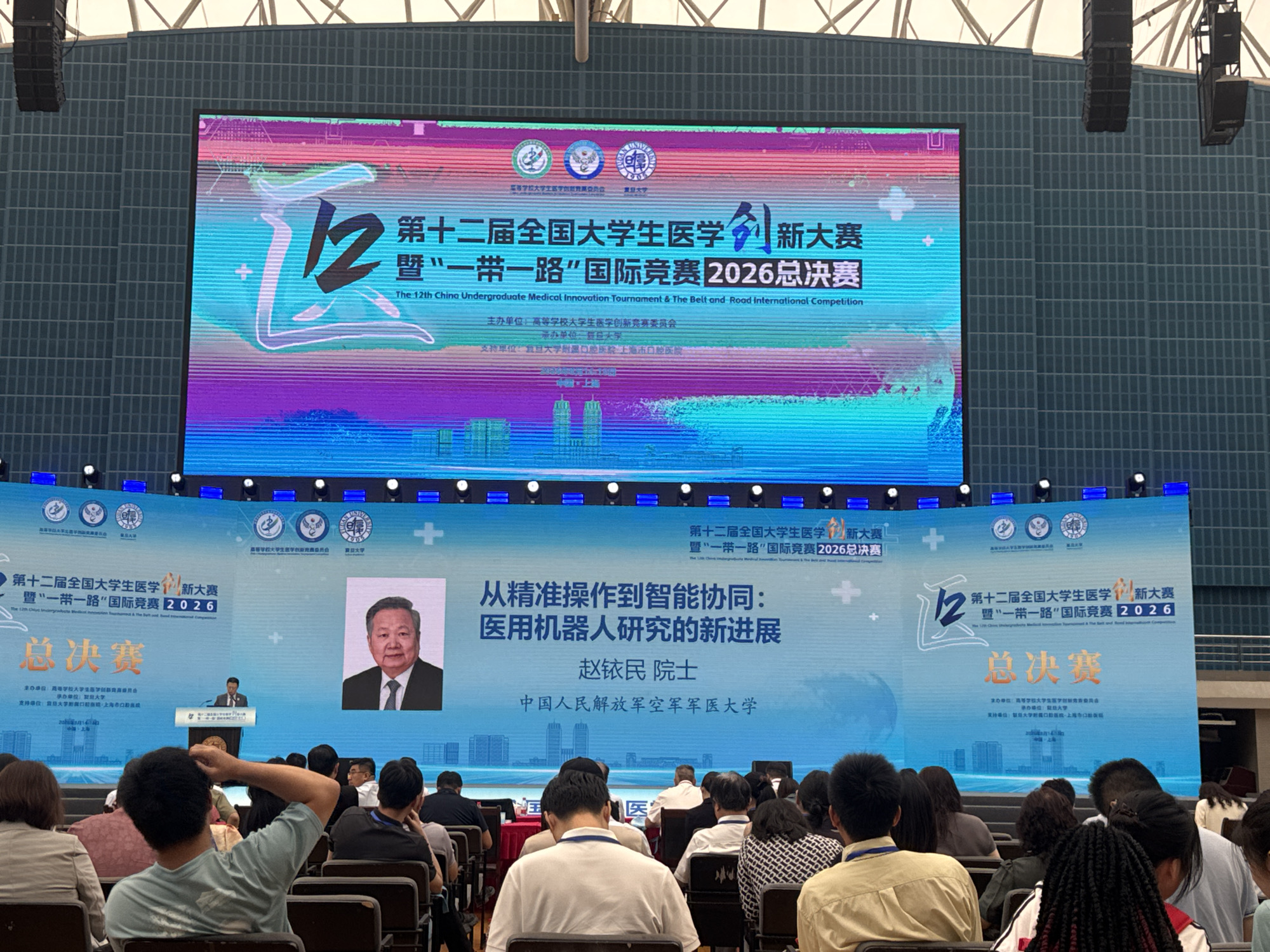 The image depicts an indoor event hall, likely a lecture hall or auditorium, with a large digital screen as the focal point. The screen displays text and graphics related to the "12th China Undergraduate Medical Innovation Tournament & The Belt and Road International Competition 2026 Finals." The screen features prominent Chinese characters and English translations, along with logos of various institutions. A portrait of an older gentleman, identified as "Zhao Minmin, Academician," is displayed on the right side of the screen, alongside the title "From Precise Operation to Intelligent Collaboration: New Progress in Medical Robot Research."
In front of the screen, on a stage, a man in a suit is seen standing at a podium, addressing the audience. The audience, composed of many individuals seated in rows of chairs, is facing the stage and the screen. The overall ambiance suggests an academic or competitive event, possibly a conference or a final round of a competition. The venue appears to be modern, with a tiled wall structure and overhead lighting. The presence of the logos and the mention of "Yangpu District, China" on the screen's information suggest that this event is taking place in Yangpu District, a district in Shanghai, China.