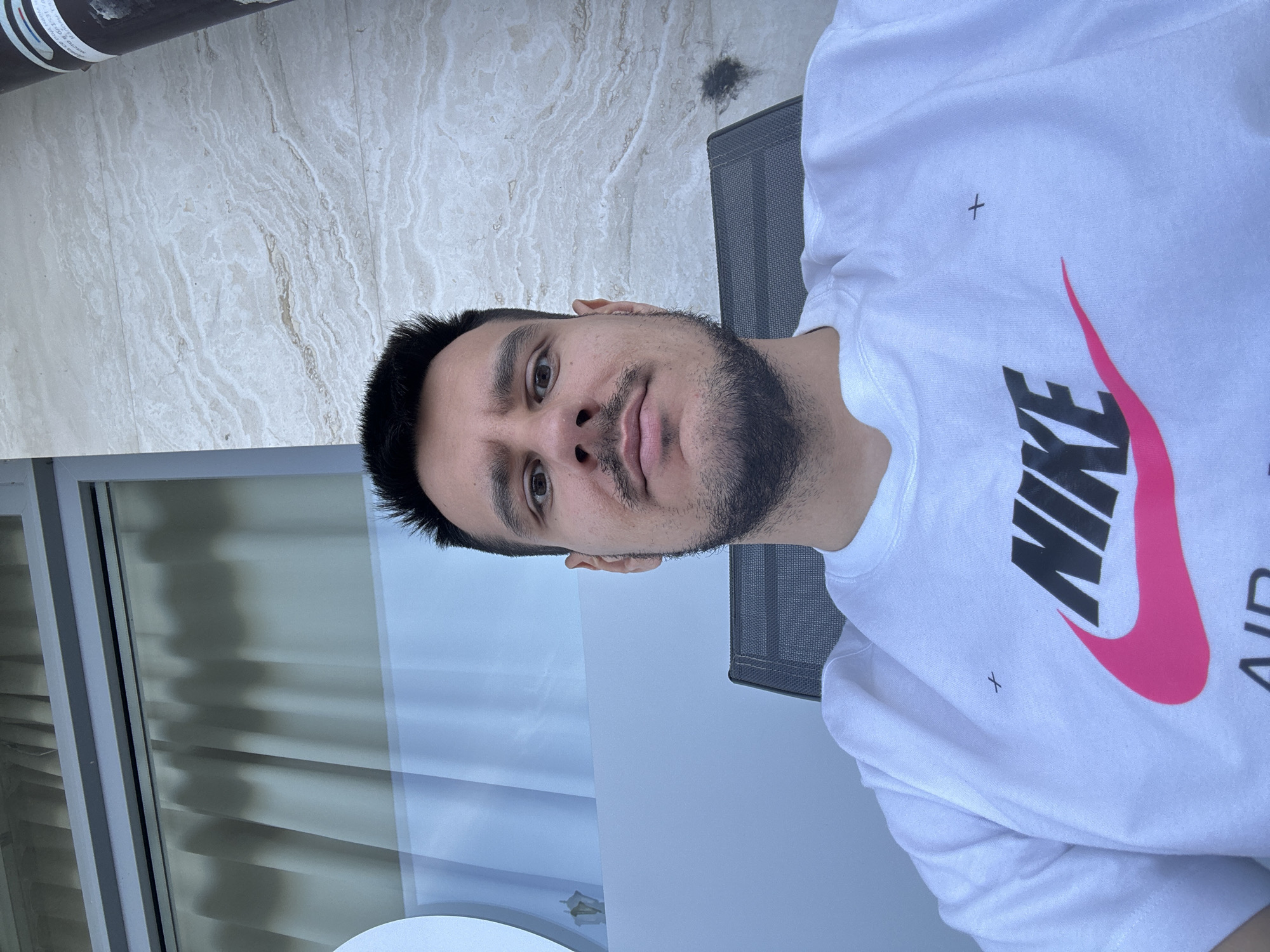 A man with dark hair and a beard is wearing a white Nike t-shirt, posing for a photograph. The background shows an exterior location with white marble walls and a large glass window with blinds. The setting appears to be a balcony or outdoor area. Without more information, a specific location within Sofia, Bulgaria, cannot be determined.