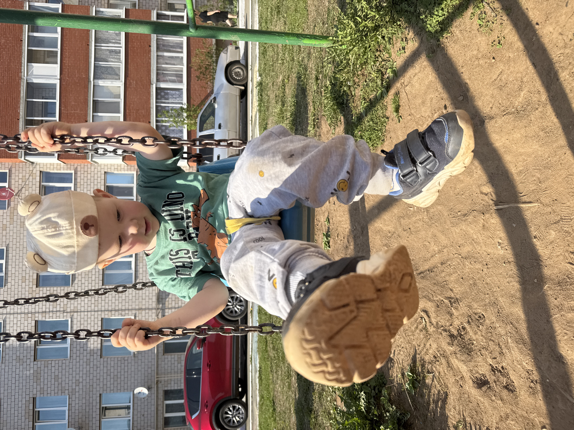 A young child, wearing a cap and green shirt, is on a swing in a playground. The child's legs are extended, showing their shoes. The playground is located in Zav'yalovo, Russia, as evidenced by buildings and vehicles typical of this location in the background. One red car and one silver car are visible behind the swing set. The ground appears to be dirt and sparse grass.