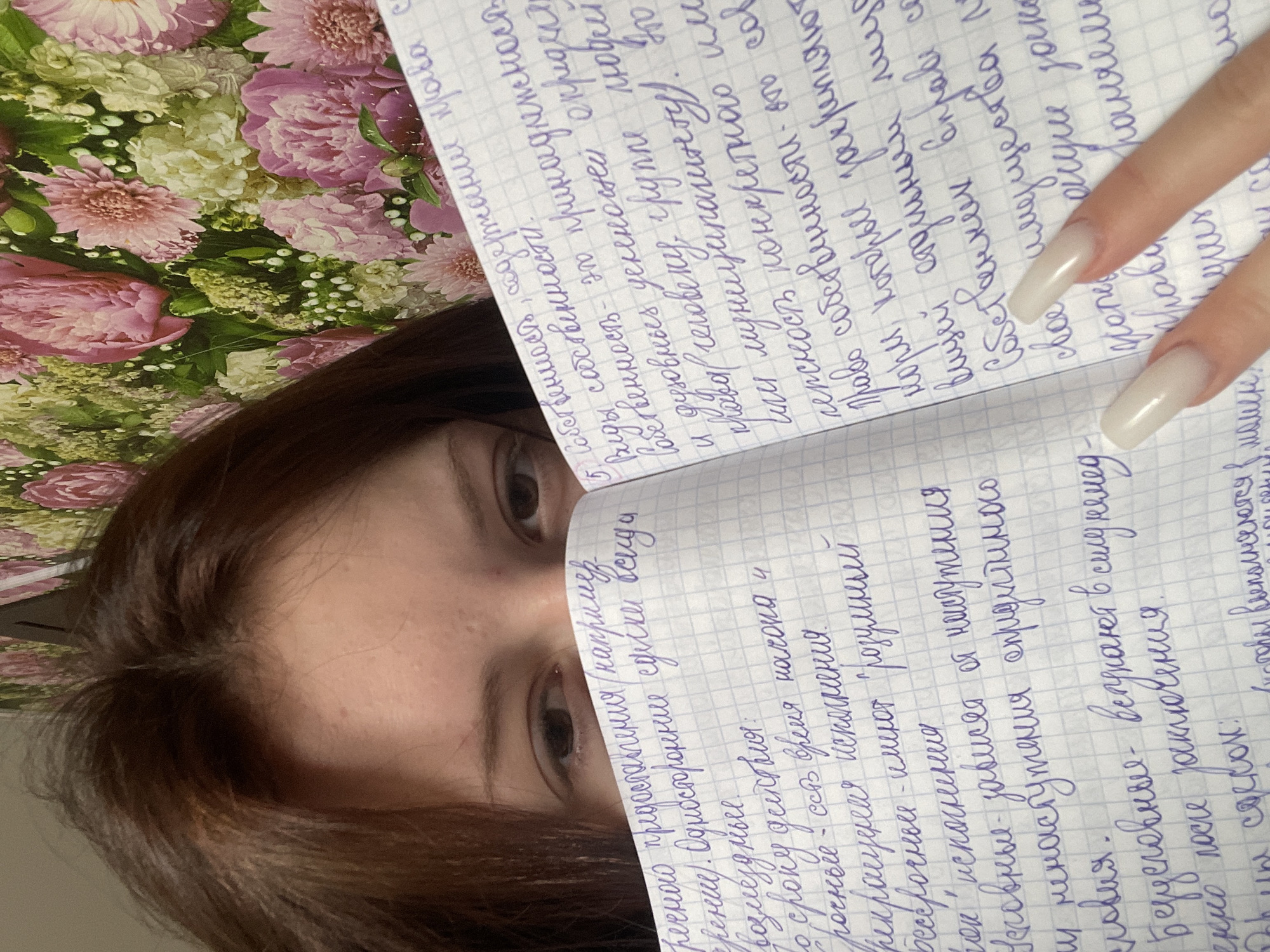The image presents a close-up, top-down view of a person whose face is partially covered by an open spiral-bound notebook. The person's eyes, part of their forehead, and dark brown hair are visible above the notebook's edge.
The notebook features grid-lined paper filled with extensive handwritten text in purple ink, using Cyrillic script. The content appears to be detailed notes or studies. On the right side of the image, a hand with visibly long, square-shaped, light-colored nails rests near the pages, indicating engagement with the notes.
The background displays a rich floral pattern composed of pink peonies, lighter blossoms, and green leaves, suggesting the notebook is placed on a decorative surface or fabric. The scene conveys a focused, indoor activity, such as studying or note-taking.