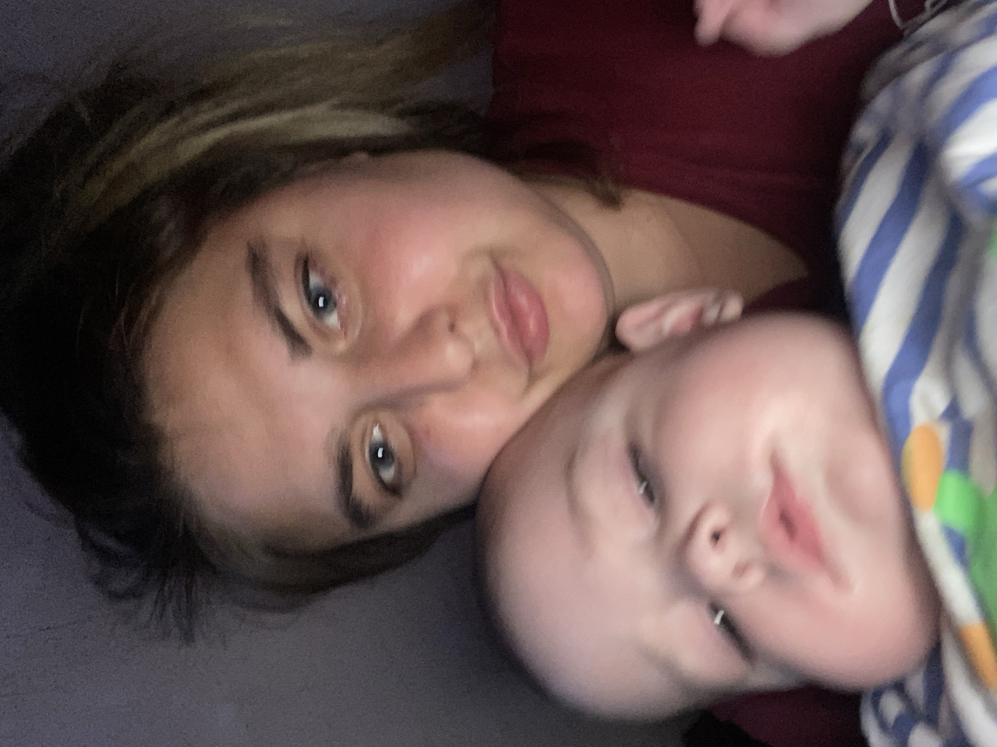 The image shows a close-up of a woman and a baby. The woman has light-colored eyes and brown hair. She is wearing a dark red shirt. The baby has light-colored skin and appears to be lying on a striped blanket. The image is dimly lit. The location is not visible in the image, and there is no information to associate this image with Otradnaya, Russia.