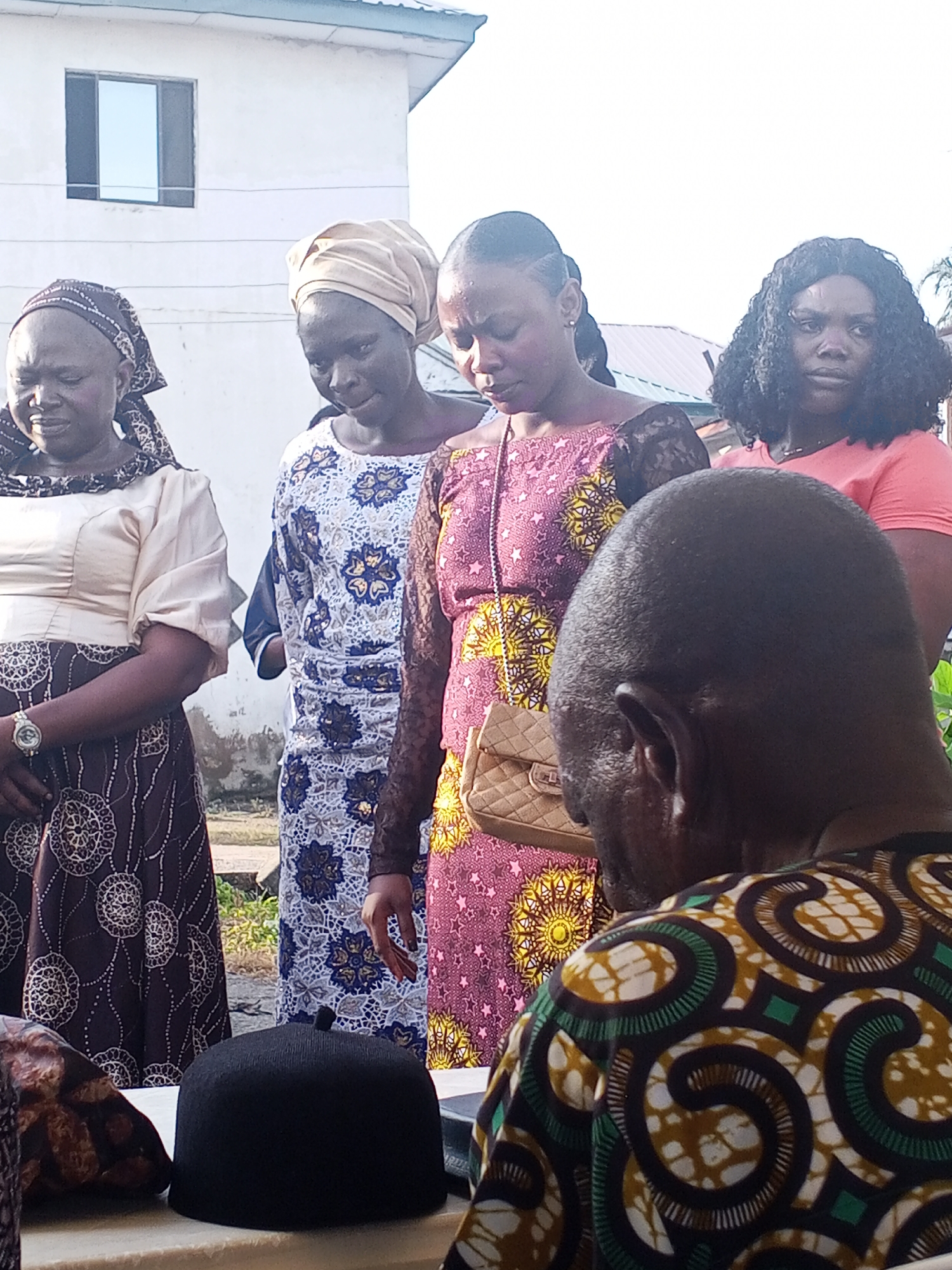 This image captures an outdoor scene in Lokoja, Nigeria, featuring a group of people, predominantly women, gathered together. The setting appears to be near a building with white walls and a section of dark-tiled roof visible. In the foreground, a man's head and shoulders are visible from behind, wearing a vibrantly patterned shirt in green, brown, and white. To his left, on a table or surface covered with a white cloth, sits a black skullcap.
The women in the image are dressed in traditional African attire. From left to right, the first woman wears a patterned headscarf and a cream-colored top with puffed sleeves, paired with a dark skirt featuring a circular pattern. Next to her, a woman in a light-colored headscarf wears a white dress with intricate blue and gold floral patterns. The third woman from the left is in the center, wearing a pink and purple patterned dress adorned with yellow floral motifs, and she carries a small, quilted beige handbag. The fourth woman, on the right, has curly black hair and wears a coral-colored t-shirt. The expressions on their faces suggest a moment of contemplation or observation, with most looking downwards. The lighting indicates it is daytime, with a bright sky suggesting fair weather. The background includes elements of the building and a hint of greenery, possibly trees, suggesting an urban or semi-urban environment within Lokoja.