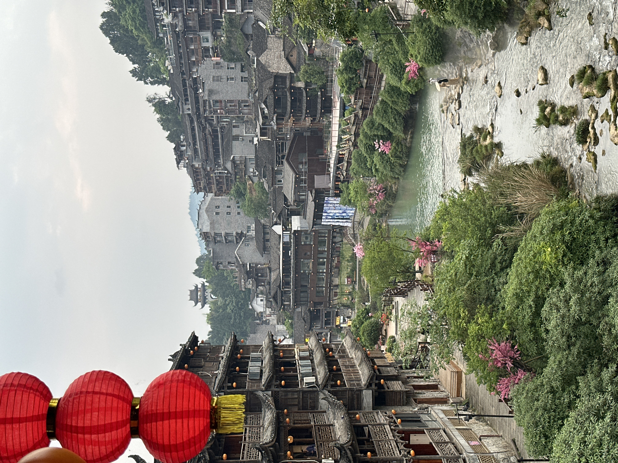 The image shows an outdoor scene in Furong, China, featuring traditional wooden buildings with intricate designs stacked along a river. Red lanterns hang prominently in the foreground. The river flows through the scene, flanked by lush greenery and trees with pink blossoms. In the background, more buildings rise up the hillside, with some structures appearing older and more weathered than others. Two individuals are seen walking in the river. The sky is overcast and appears foggy.
