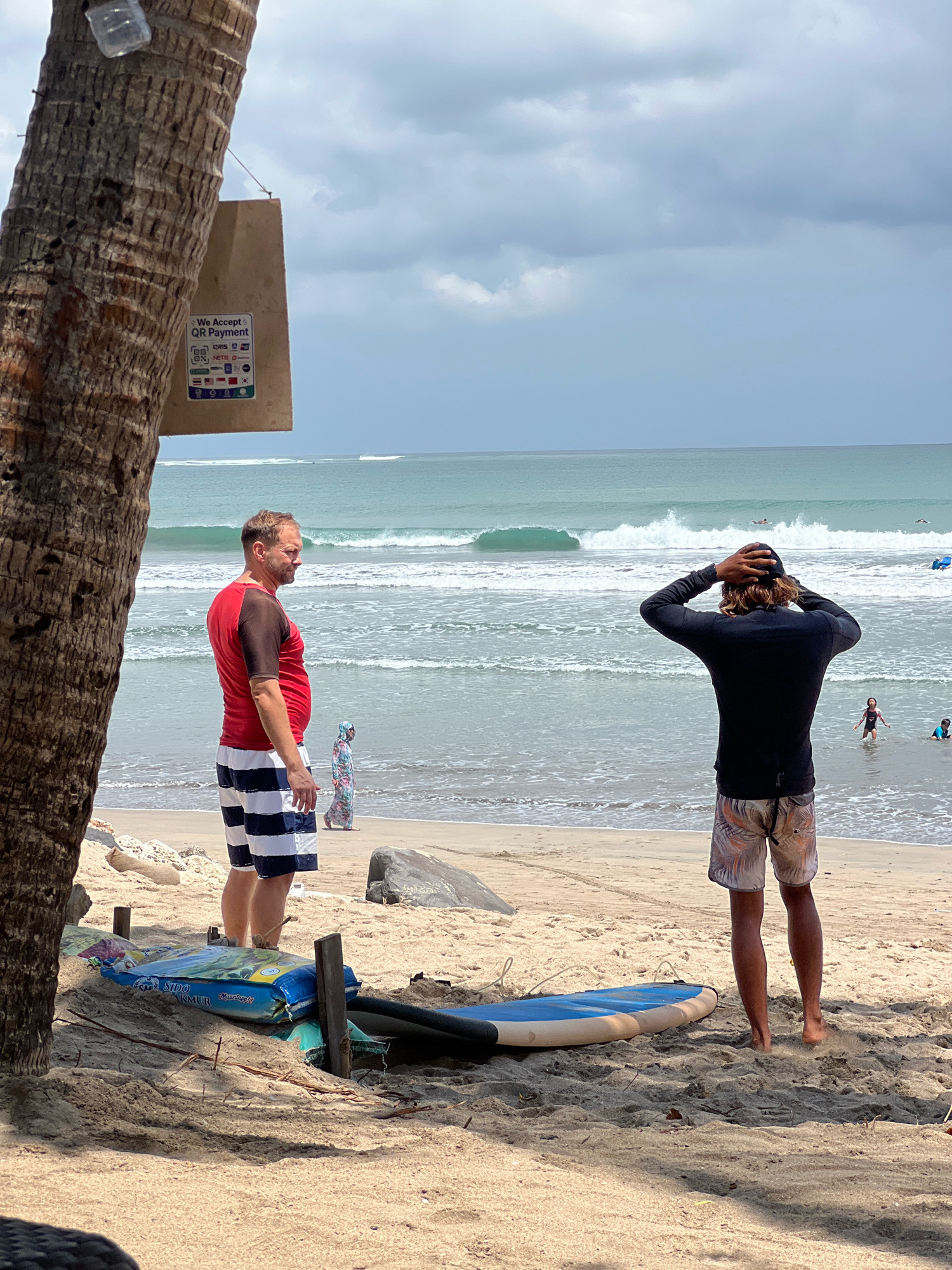 This is a medium shot image taken at Kuta Beach, Indonesia, focusing on the shoreline and the ocean. In the foreground, partially obscured by a palm tree trunk, are two men facing away from the camera, looking out at the ocean. The man on the left wears a red and black t-shirt and blue and white striped shorts. The man on the right wears a black long-sleeved shirt and patterned shorts. In front of the men are two surfboards lying on the sand, one blue and one with a blue and yellow design.
The beach is sandy with some rocks. The ocean has moderate waves, with several surfers visible in the water. Further back on the beach, a woman in a floral-patterned dress walks away from the camera. The sky is overcast with scattered clouds. A sign is visible attached to the palm tree, indicating that QR payments are accepted. The overall scene depicts a typical day at a popular Indonesian beach, with people enjoying the water and the scenery.