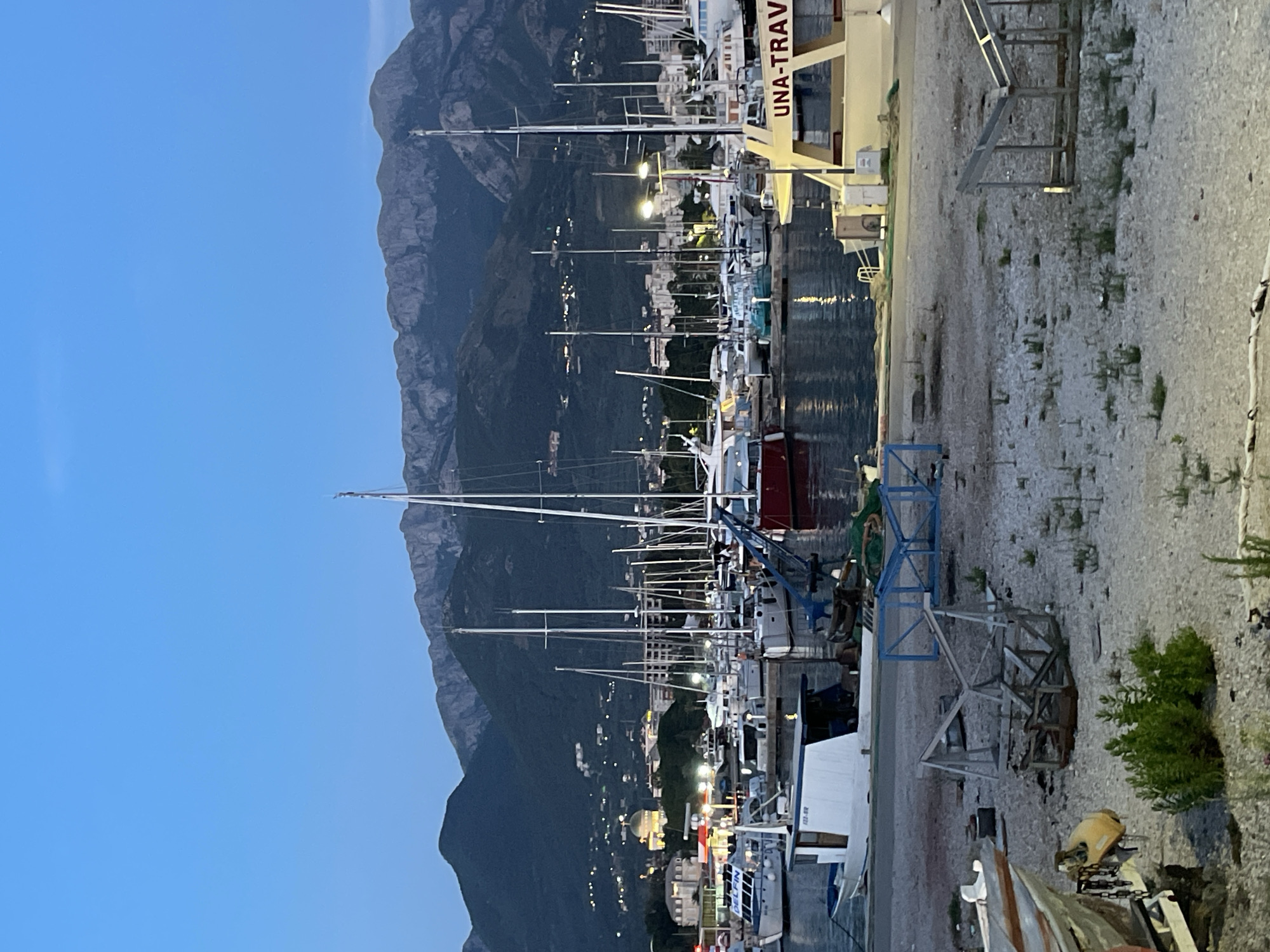 The image depicts a harbor scene in the city of Bar, Montenegro, at dusk. The sky is a deep twilight blue, transitioning from a lighter hue at the horizon. In the background, a range of mountains is visible, with lights from buildings scattered across their lower slopes. The foreground is dominated by a harbor filled with numerous sailboats and smaller motorboats moored together. Many of these boats have tall masts, creating a dense forest of rigging.
On the right side of the image, a yellow and white boat with "UNA-TRAV" written on its side is prominently featured. To the left of this boat, a white and blue motorboat with the markings "623-BR" is docked. In the immediate foreground, the ground is covered in gravel and scattered debris, including pieces of wood and what appears to be a boat trailer or cradle. Some green vegetation is growing amongst the gravel. The water in the harbor reflects the lights from the boats and the distant shore. There are no people clearly visible in the image. The overall atmosphere is serene and quiet, characteristic of a harbor at the end of the day.
