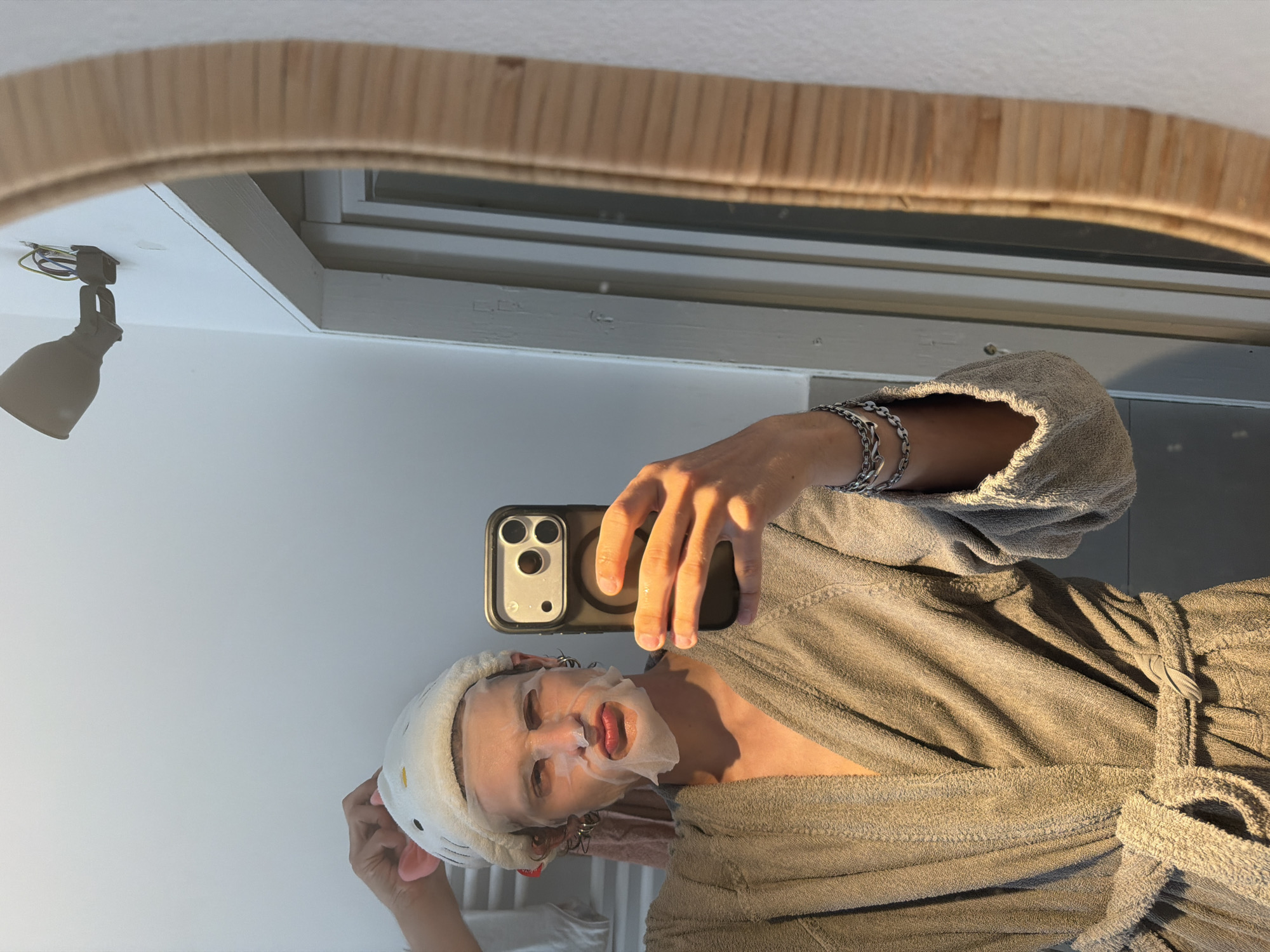 A person wearing a gray robe, a face mask, and a headband with cat ears is taking a selfie in a mirror. The mirror has a light wooden frame. Reflected in the mirror are a light fixture, a window, and white walls. The person is holding a smartphone with three camera lenses. The environment suggests an indoor setting, possibly a bathroom. The scene does not provide enough information to determine the location within the city of Milan, Italy.