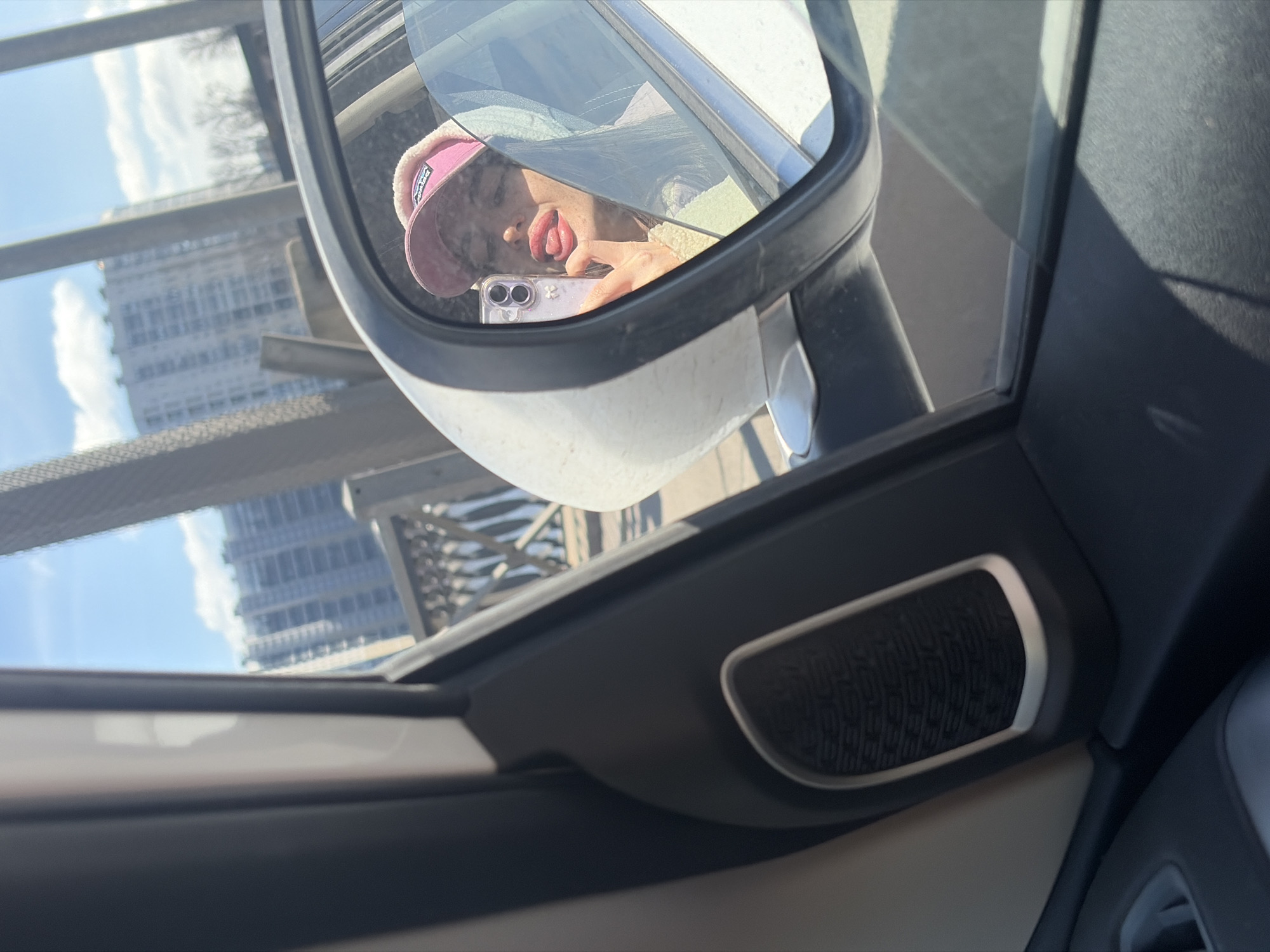 The image shows a rearview mirror reflecting a young person with a pink hat, sticking out their tongue while holding a smartphone. In the background, a building is visible, suggesting an urban setting. Based on the architectural style of the building in the mirror and the atmospheric conditions (clear sky with some clouds), the location may be Saint Petersburg, Russia. The photo is taken from inside a vehicle, with the car's door panel and speaker visible in the foreground.