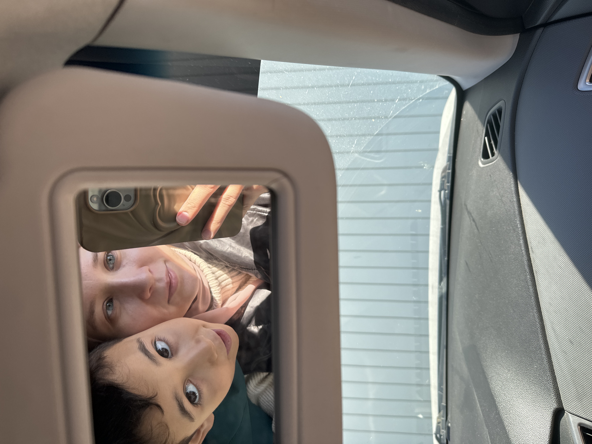 A woman and a child are visible in the mirror on the sun visor of a car. The woman is holding a phone, presumably taking the picture. A gray dashboard with an air vent is visible to the right of the mirror. The exterior of the car window shows a light-colored, possibly snowy, landscape or building. The exact location within Chaikovsky, Russia, cannot be determined from the image.
