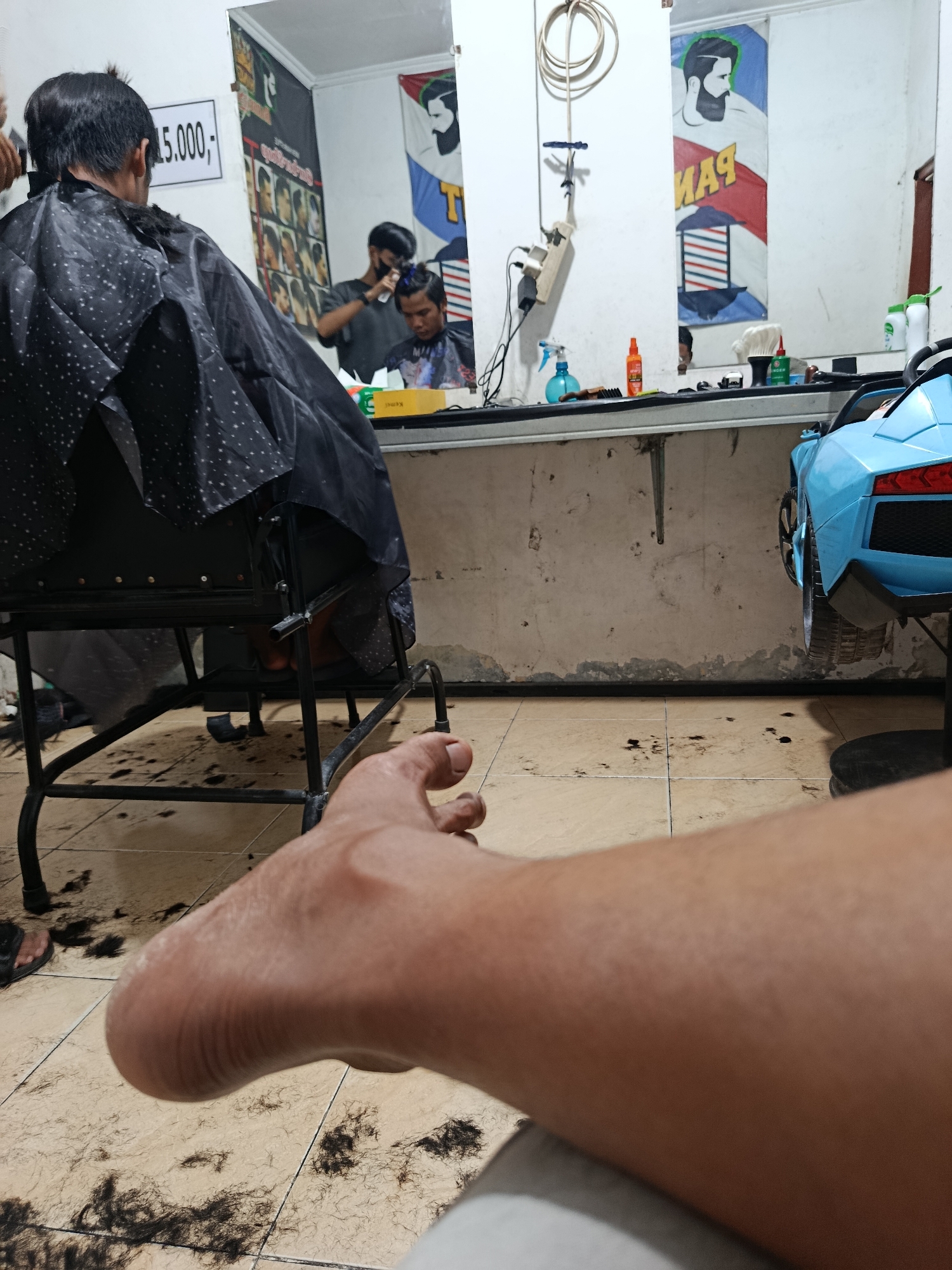 The image depicts the interior of a barbershop, likely located in Sarwadadi, Indonesia, as indicated by the currency notation "15.000,-" on a sign. The foreground is dominated by a close-up of a person's leg and foot, resting on the tiled floor which is scattered with numerous dark hair clippings. Behind the leg, a black barber's chair is visible, draped with a patterned cape. The midground reveals a barber at work, reflected in a large mirror that covers a significant portion of the back wall. In the reflection, a barber wearing a mask is attending to a seated customer who is also wearing a cape. Another barber is visible in the background of the reflection, working on a different customer. The mirror also displays posters with barber-related imagery. The environment appears functional, with barbering tools and supplies visible on a counter. A toy ride-on car, possibly a child's prop, is partially visible on the right side of the frame. The lighting suggests an indoor setting, possibly artificial.