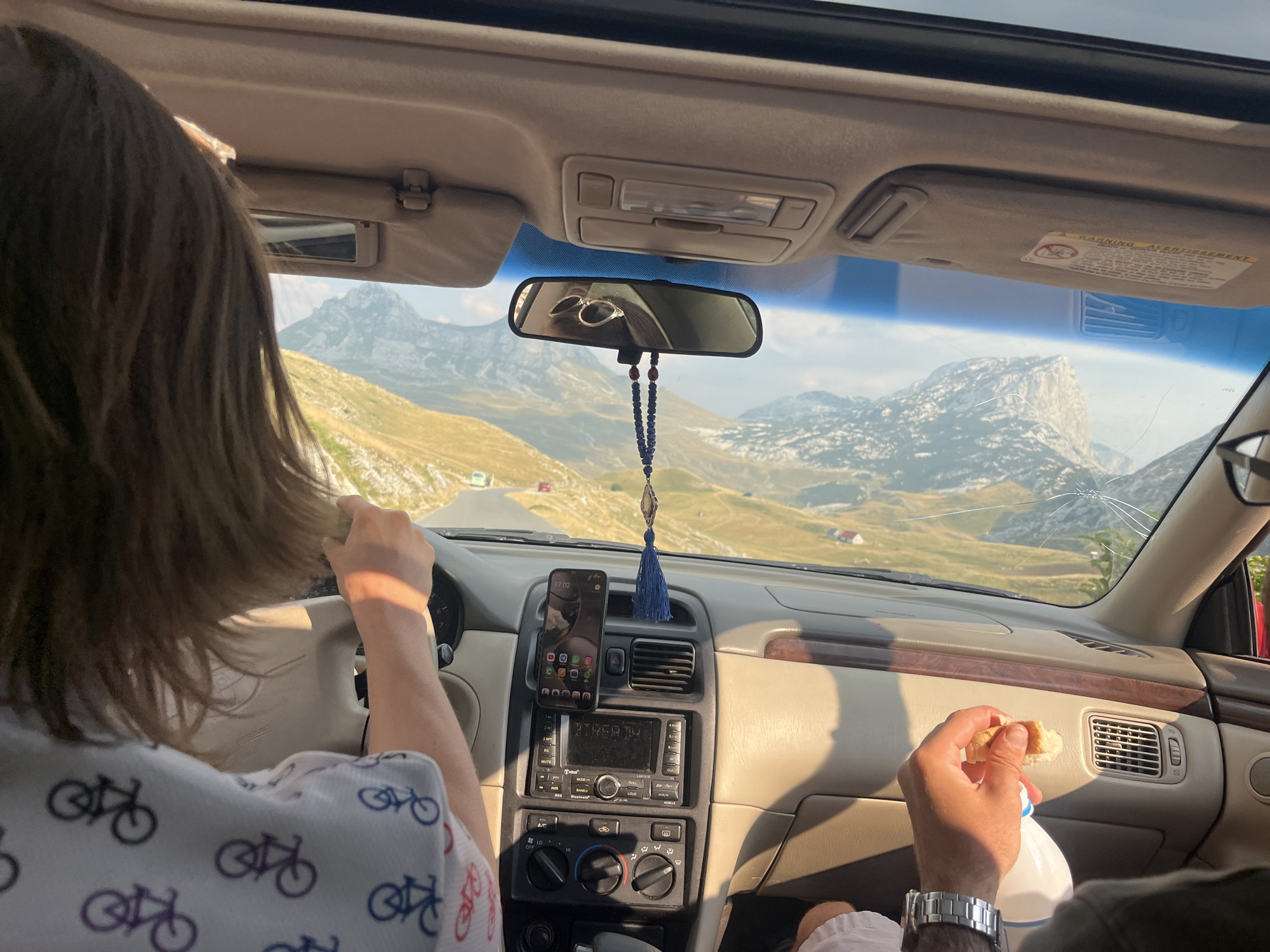 The image shows the interior of a car traveling on a road in the mountainous landscape of Dubrovniksko, Montenegro. The perspective is from the passenger seat, looking forward through the windshield and towards the driver. The driver, with shoulder-length brown hair, is seen from behind, with their left hand on the steering wheel and their right hand touching their hair. They are wearing a white shirt with a repeating pattern of bicycles.
The car's dashboard is visible, featuring a smartphone mounted on the air vent displaying the time as 17:02 and various app icons. Below the phone is the car's audio system. A blue beaded tassel hangs from the rearview mirror. In the passenger's side of the car, a hand is holding a piece of bread, seemingly about to eat it, next to a bottle of water. The hand is wearing a silver wristwatch.
The windshield shows a scenic view of rolling hills and imposing mountains under a clear sky. A winding road is visible ahead, with a few other vehicles in the distance. The overall impression is of a journey through a picturesque, rural, mountainous region. The sun appears to be setting or is in the late afternoon, casting a warm light on the scene.