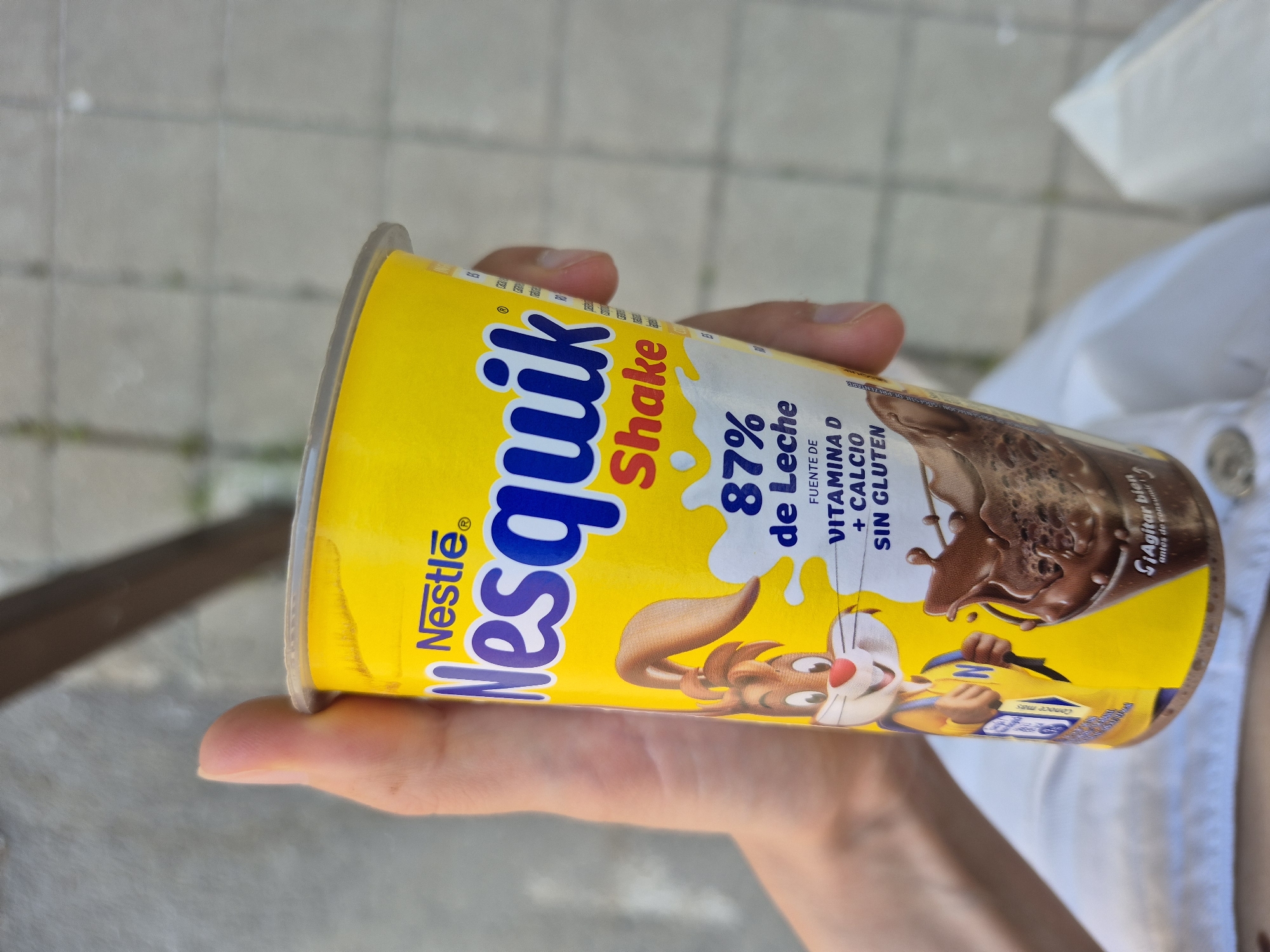 A person holds a yellow Nesquik Shake beverage container. The container has blue and red text that reads "Nesquik Shake." It also features graphics of a rabbit character and chocolate splashes. Text on the container includes "87% de Leche," "FUENTE DE VITAMINA D + CALCIO SIN GLUTEN," and "Agitar bien antes de consumir." The person's hands are visible holding the container. The background appears to be an outdoor setting with a paved surface. The lighting suggests it is daytime.
