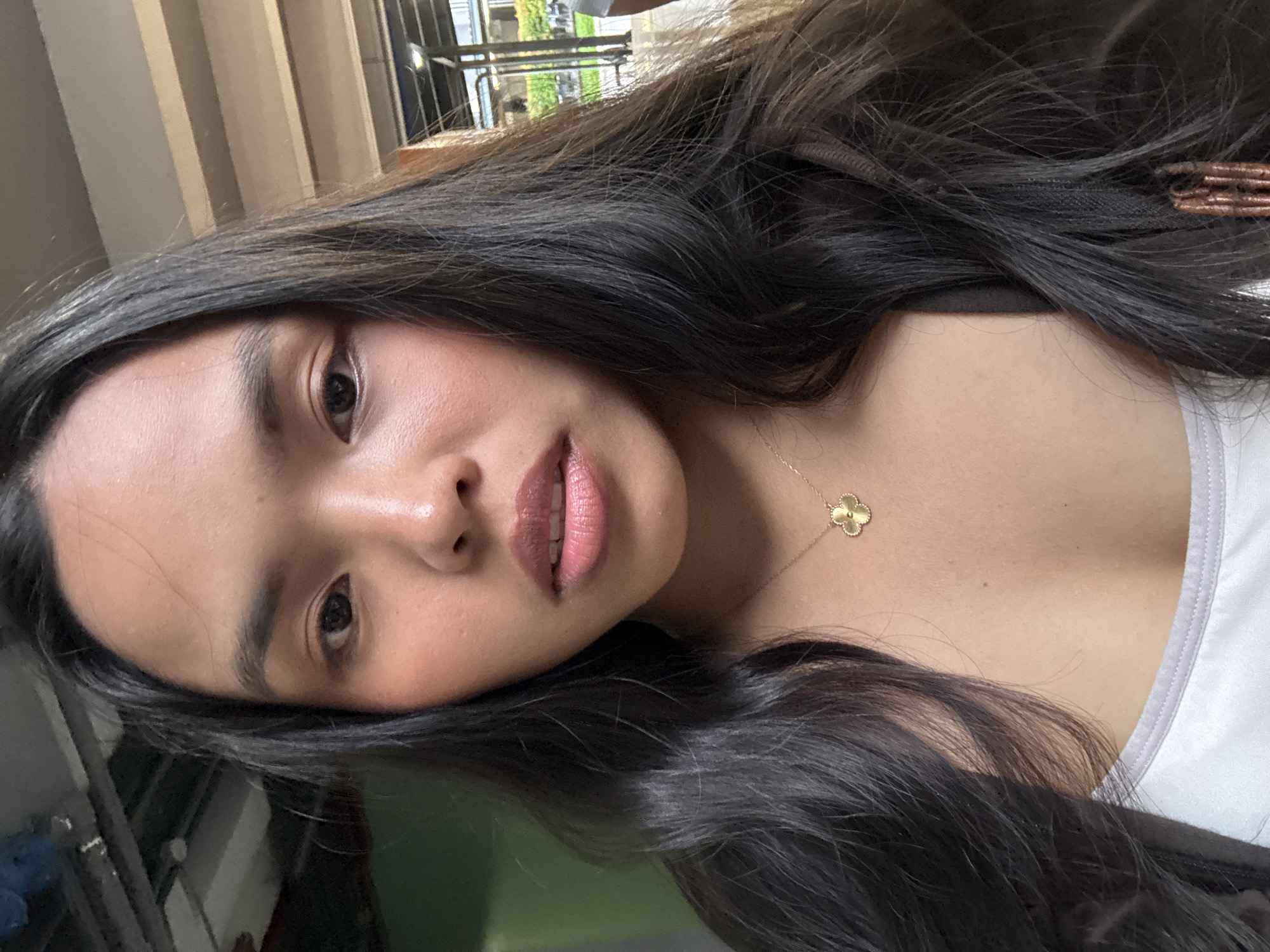 A young woman with long, dark hair is captured in a close-up, frontal selfie. She is wearing a white tank top with a grey trim and a delicate gold necklace with a clover-shaped pendant. Her makeup includes blush on her cheeks and lip gloss. The background appears to be an indoor or covered outdoor space with industrial-style lighting and what could be ceiling beams visible above. To the right, there is a glimpse of an outdoor area with greenery and a building.
While the image itself does not contain specific markers to pinpoint its exact location within Davao City, Philippines, the overall aesthetic of the lighting and surrounding architecture suggests a common urban setting, possibly a mall, transportation hub, or a commercial establishment found in such a city.
