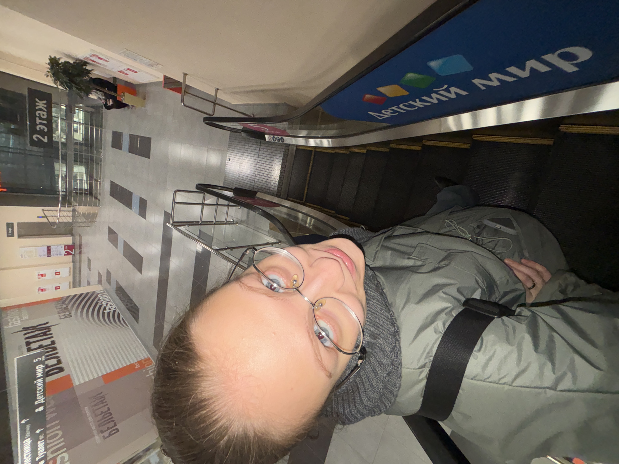 A top-down, distorted perspective photograph shows a person riding an escalator in what appears to be a shopping mall. The person, who has fair skin and is wearing glasses, a gray turtleneck sweater, and a green jacket with a black strap, is looking directly into the camera. The escalator has a blue and green sign in Russian reading "Детский мир" (Detsky Mir), which translates to "Children's World". The surrounding environment shows the mall's interior, including signage and flooring, with the words "БЕЛЬЕТАЖ" (Belyetaj), which is a reference to the fashion store located on the second floor. In the background on the left side, there is signage indicating the location of "Детский мир 5". The location of this photo is in the city of Perm, Russia.