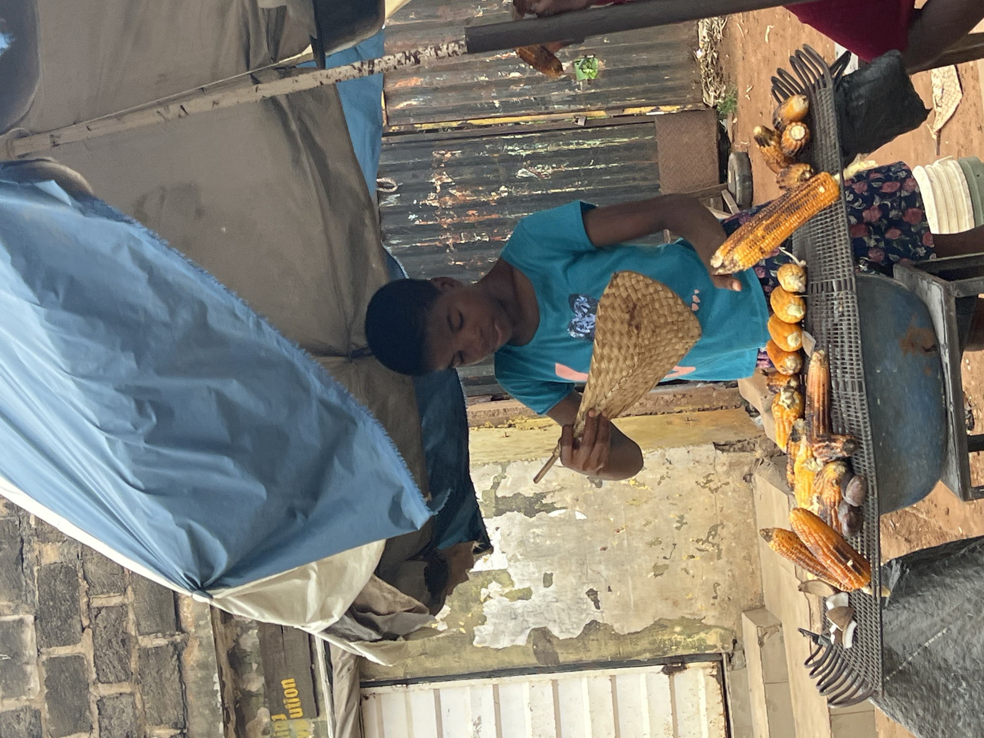 A young individual stands under a blue and beige tarped canopy at a street-side vendor stall in Nkpor, Nigeria. The individual, wearing a turquoise t-shirt and patterned shorts, is fanning a woven mat over a grill laden with several ears of grilled corn. The stall is set up in front of a weathered brick building, with a corrugated metal fence in the background. The ground is dirt and scattered debris. Other items on the grill include what appear to be small, rounded fruits or vegetables and possibly pieces of coconut. The overall scene depicts a typical market or roadside food selling operation.