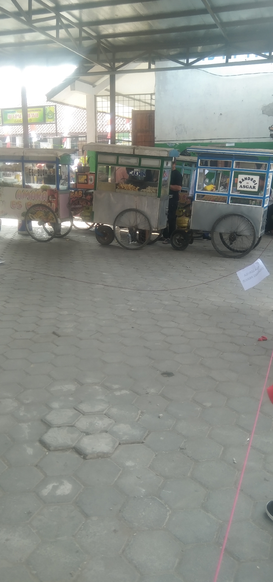 In Rahayu, Indonesia, an outdoor scene unfolds under a covered structure with a metal roof. Two food carts, each with wheels and awnings, are parked side-by-side on a hexagonal paver surface. The cart on the left displays "ES BUAH" and "CAMARA ES CIEM," suggesting it sells fruit ice and possibly ice cream. The cart on the right has signage for "CAKMIN" and "BANDROS ASGAR." A person in a black shirt stands near the right cart, next to a yellow gas tank. The ground is paved with grey, hexagonal interlocking stones. A white sign with black text, possibly a notice or award, is placed on the ground in the lower right quadrant of the image, with some red objects visible nearby.