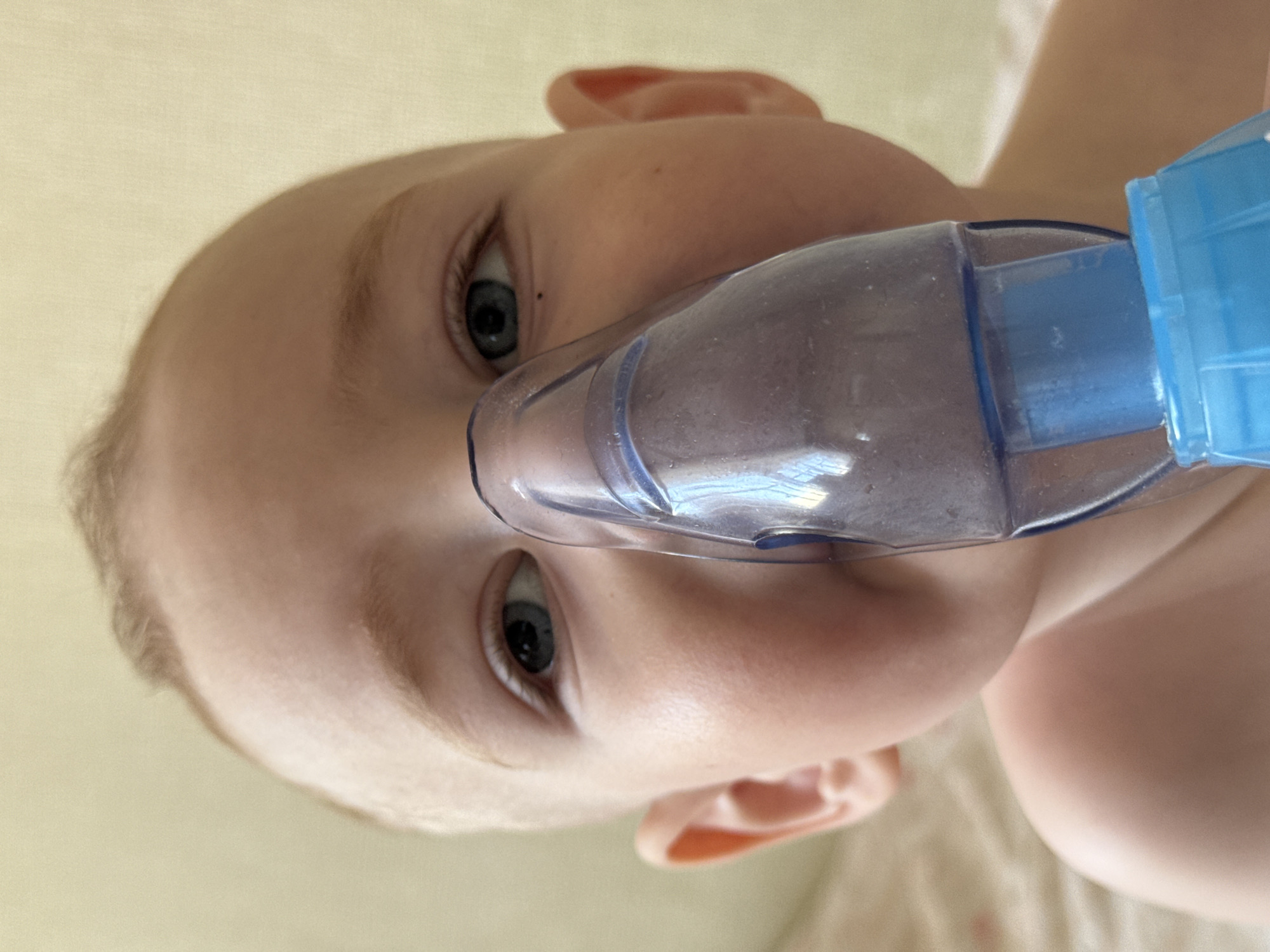 A close-up, tilted shot depicts a fair-skinned child wearing a blue and clear nebulizer mask. The child's face, dominated by large, gray-blue eyes, fills most of the frame. The background appears to be a light beige surface, likely a bed sheet or other soft fabric. The location in Chaikovsky, Russia, is unknown.