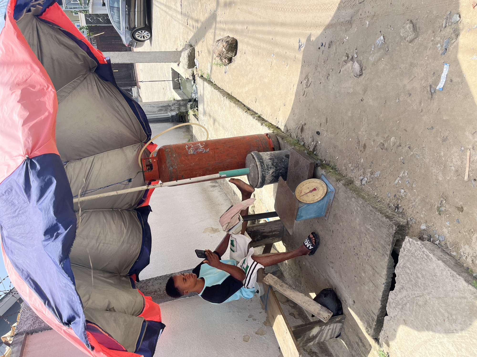 A man is sitting under a makeshift shelter in Iranla, Nigeria, looking at his phone. The shelter has a blue, pink, and gray tarp covering it. Next to him is a red propane tank, a black cylinder, and a blue scale. The area is paved with concrete. The ground is cracked and broken in places. A car is parked nearby.