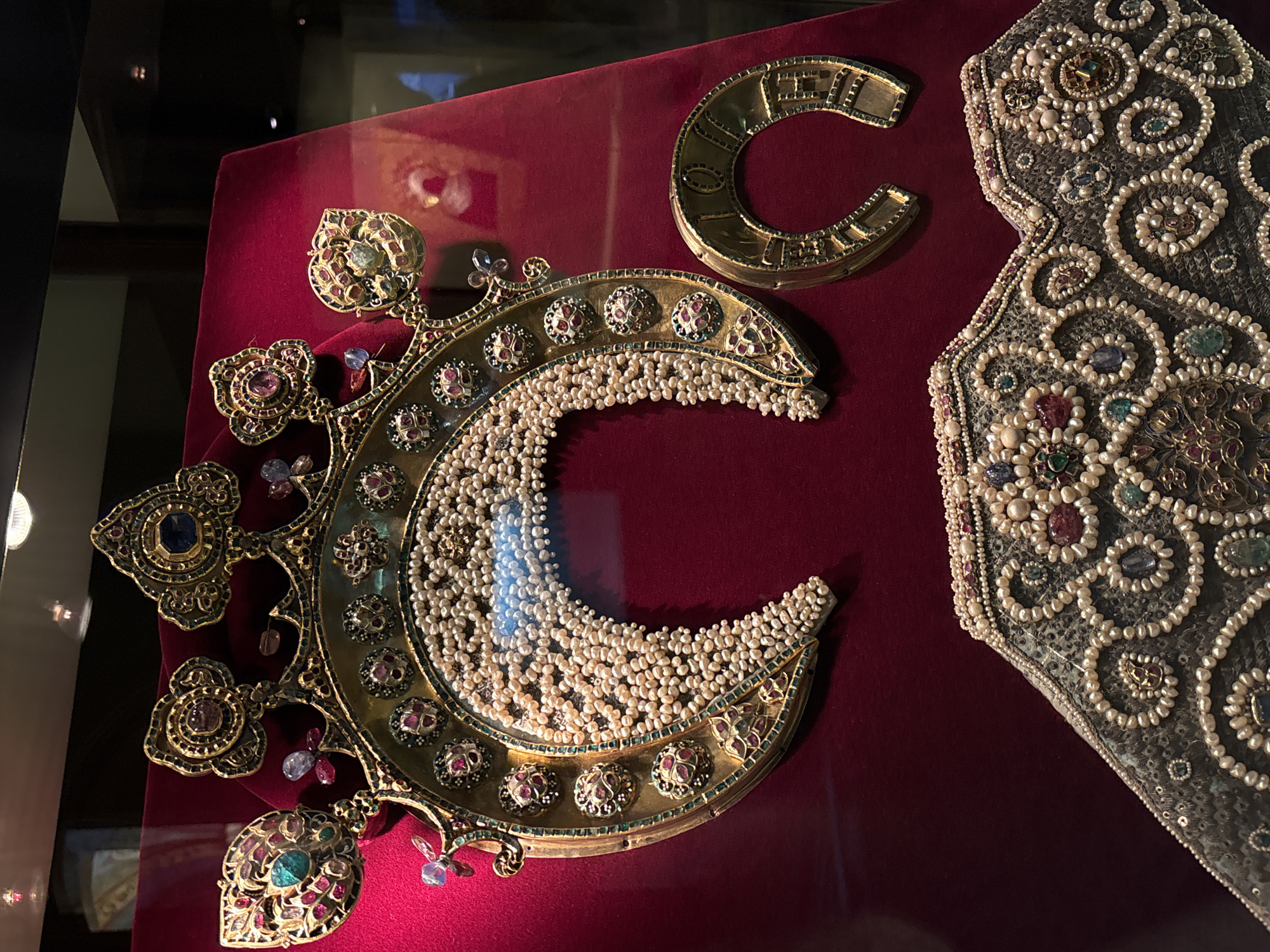 The image shows several ornate, historic items displayed on a red surface, likely within a museum setting. One prominent item is a large, crescent-shaped piece adorned with pearls and colorful gemstones. To the right of this, there is a fragment of material, also heavily decorated with pearls and gemstones in an elaborate swirling pattern. Above the crescent, there is a smaller, crescent-shaped golden element, possibly part of a larger piece. Given the intricate detailing and precious materials, these items are likely historical artifacts of significant cultural or historical value. While the location is noted as Moscow, Russia, the image alone does not confirm it. The objects appear to be behind glass, indicating a museum or exhibition display.
