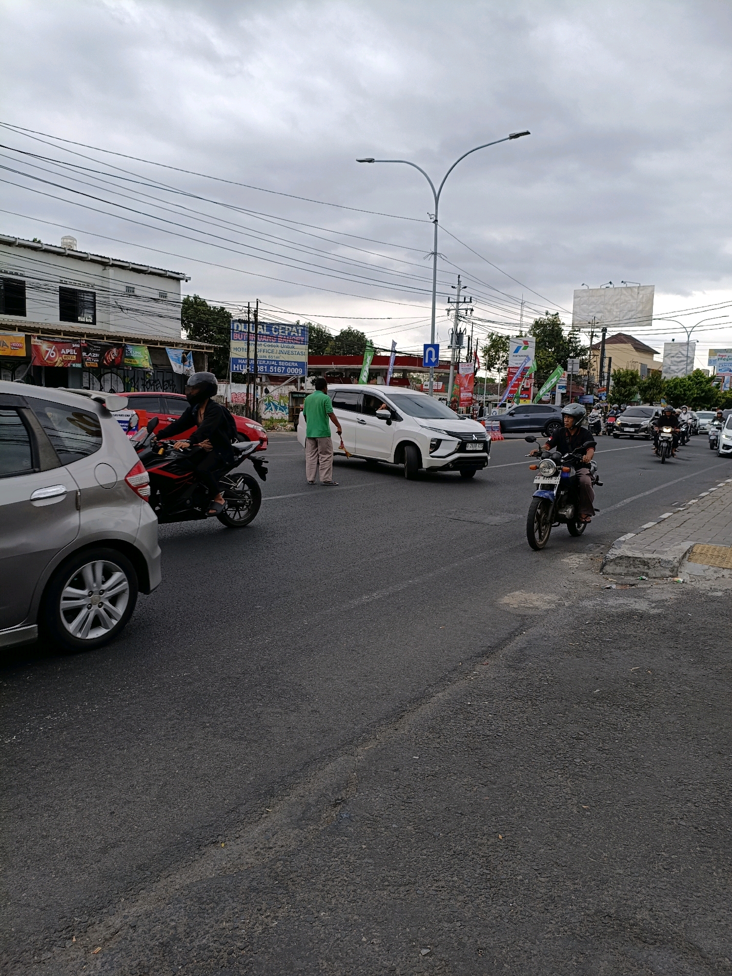 The image depicts a street scene in Depok, Indonesia, likely during daylight hours with overcast skies. The foreground shows a gray car in motion on an asphalt road with visible cracks and patches. To the left, a motorcycle rider wearing a black jacket and helmet is also moving. In the middle ground, a white SUV is stopped at an intersection, and a pedestrian in a green shirt and light-colored pants is crossing the street, holding a yellow object. Further back, a blue motorcycle is being ridden by a person wearing a helmet and dark clothing. The background features buildings, streetlights, power lines, and several billboards. One prominent billboard displays text in Indonesian, including "DIJUAL CEPAT" (For Sale Quickly). The overall scene suggests a moderately busy urban environment with traffic and pedestrians.