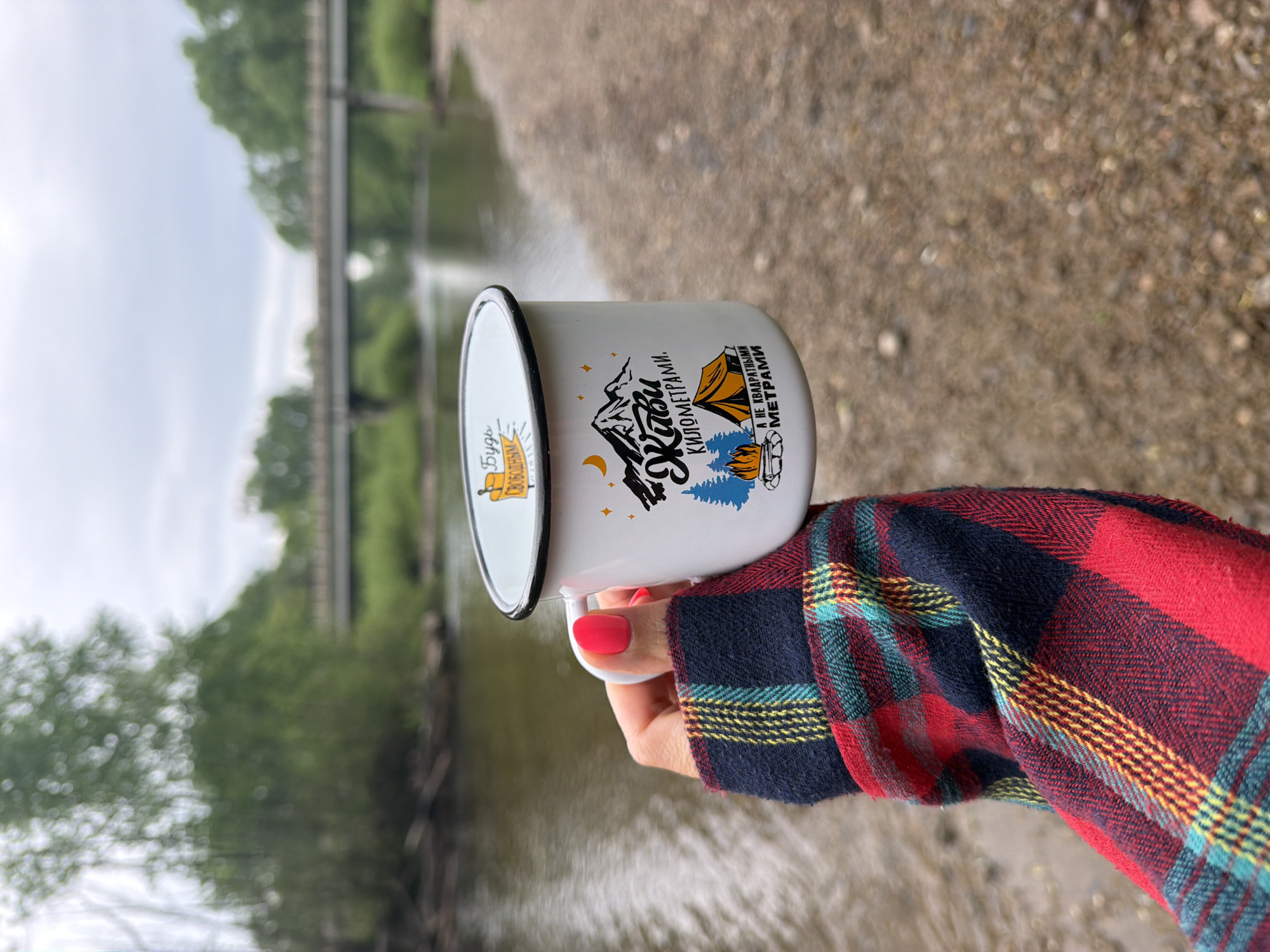 A person is holding a white enamel mug with a black rim in front of an outdoor scene. The mug has two designs on it. The first is a yellow circle with text in Cyrillic script. The second is a more complex design with mountains, a tent, trees, and stars. The text on this design is also in Cyrillic. The person is wearing a red and black plaid shirt. The background includes a body of water, a bridge or overpass, and trees. The photograph was taken in Trek, Russia.