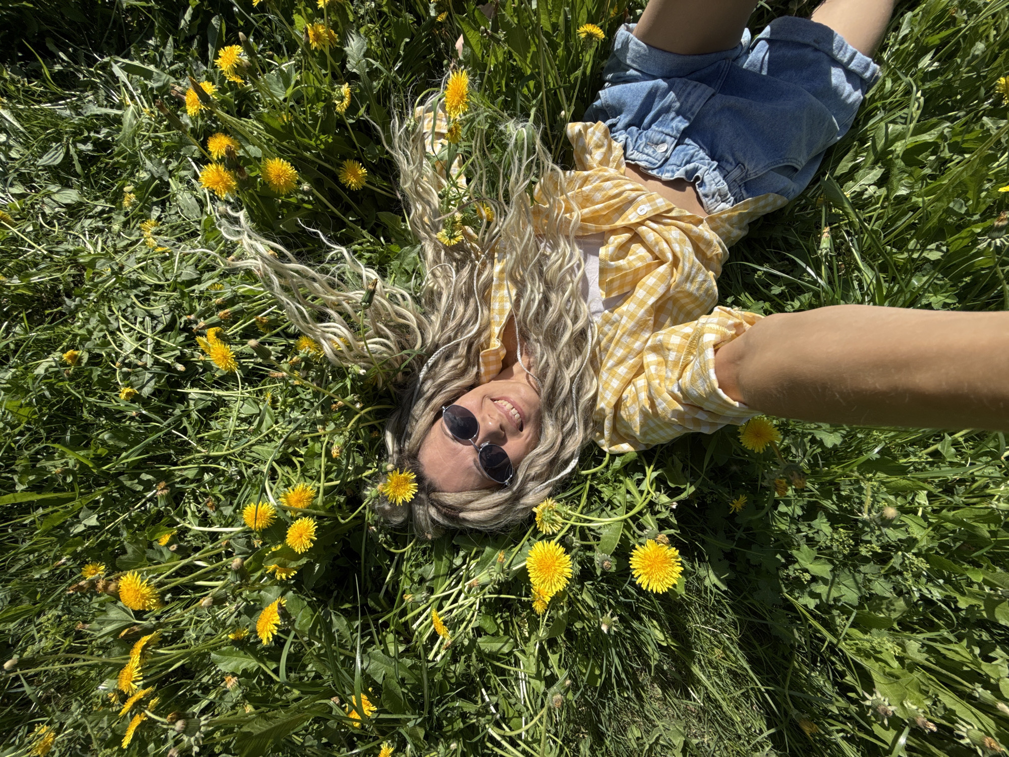 A woman lies in a field of dandelions. She wears denim shorts, a white tank top, and a yellow and white checkered shirt. Her blonde hair is styled in thick braids and she wears round sunglasses. Her right arm extends towards the camera. The background is a grassy field dotted with bright yellow dandelions. This is likely a photograph taken in Rusanovo, Russia.