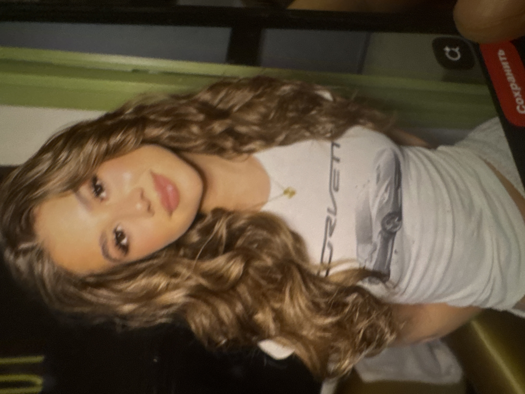 A photograph captures a digital screen displaying a young woman. The woman has long, voluminous wavy/curly light brown hair and appears to be smiling subtly. She is wearing a white t-shirt featuring a graphic print of a vintage car, likely a Corvette, with the partial text "ET" visible. A delicate gold chain necklace with a small pendant rests around her neck. The image on the screen is rotated, with her head tilted to the left. In the top right corner of the digital display, a camera rotation icon is visible. The physical device displaying the image has a finger partially visible in the upper right, holding it. A red bar is also present on the right edge of the physical device, with the Russian word "Сохранить" (Sokhranit', meaning "Save") printed in white text. The background behind the digital device includes a horizontal green element. The overall scene suggests a casual portrait or selfie being viewed or saved on a mobile device.
