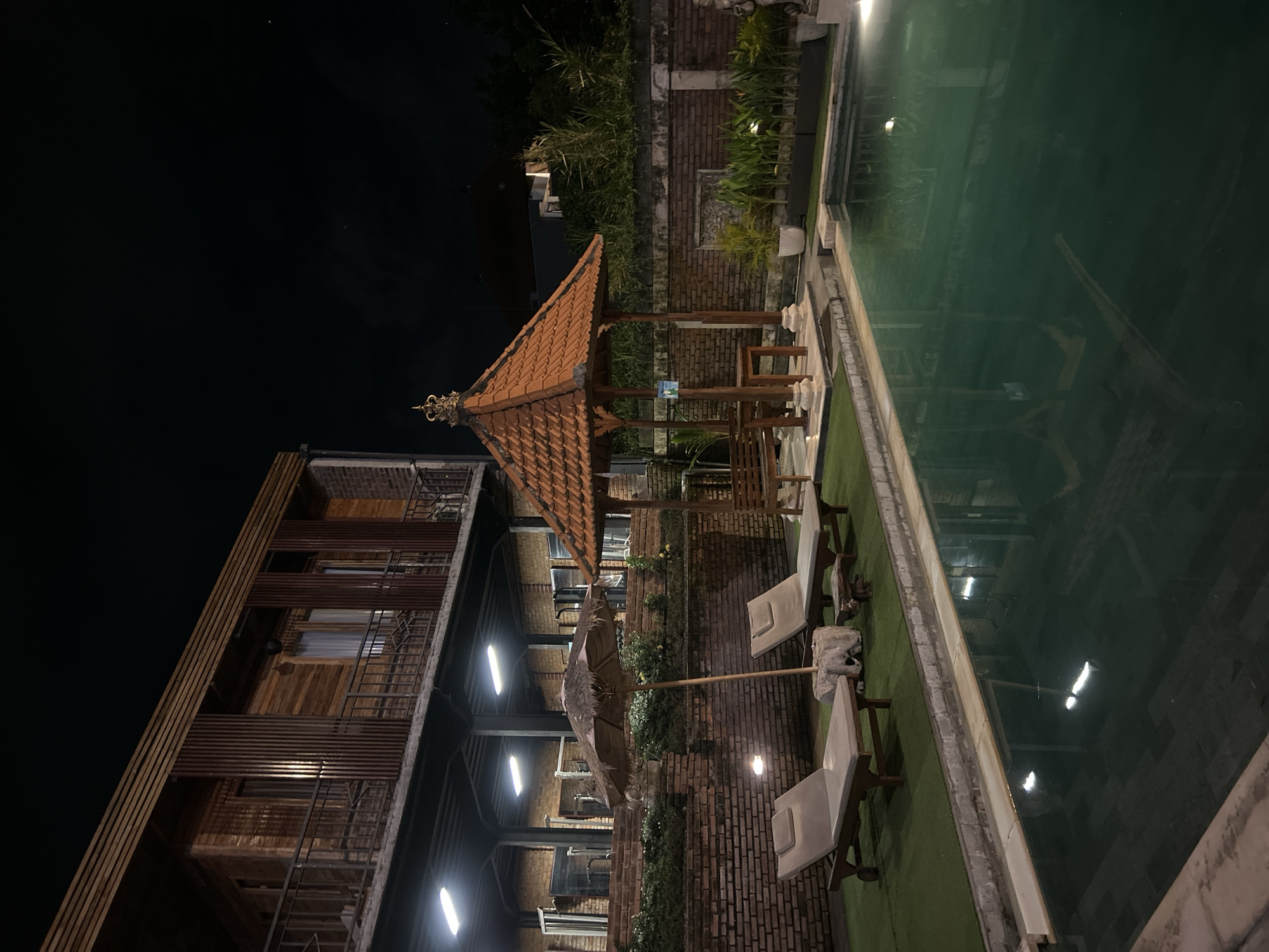 A nighttime view of a resort pool area in North Kuta, Indonesia, shows a multi-story wooden building with balconies, a traditional Balinese-style gazebo with a terracotta tile roof, two lounge chairs with umbrellas, and a swimming pool. The building's facade is clad in horizontal wooden planks and vertical slats, with balconies and railings visible. The gazebo, centrally located, features a decorative finial on its roof. Two light-colored lounge chairs are placed on a patch of artificial turf near the pool. The swimming pool, filled with dark green water, reflects the lights from the surrounding area. A brick wall borders the property, and potted plants are arranged along it. The sky is dark, indicating nighttime.