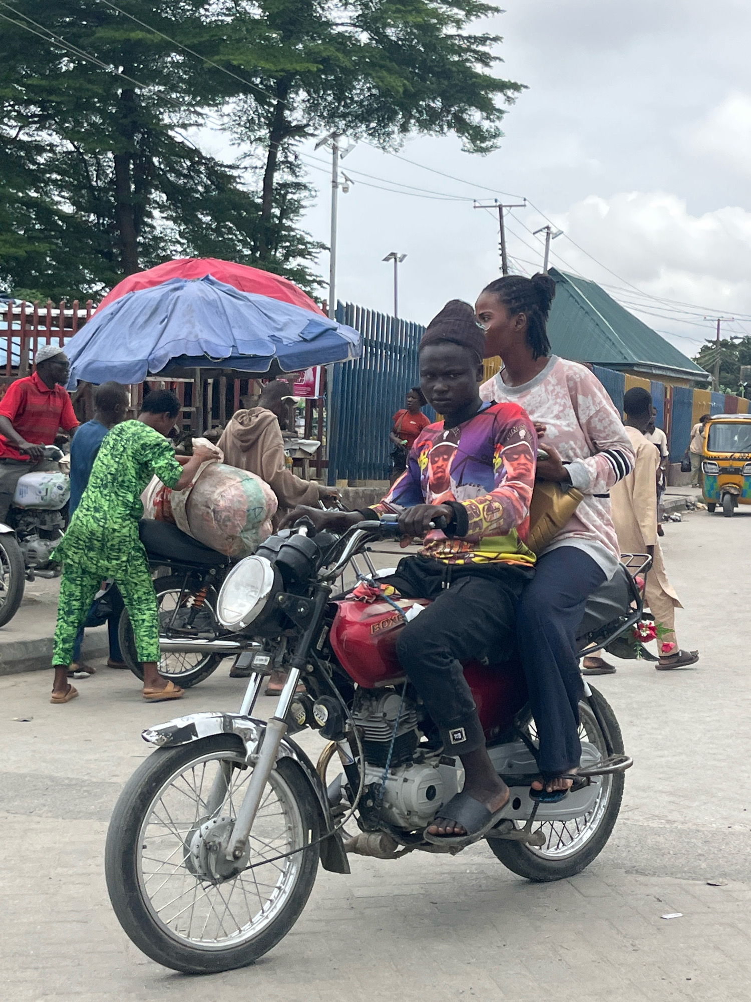 In Ado, Nigeria, a young man wearing a colorful, patterned long-sleeved shirt and black pants rides a red motorcycle. He is wearing dark sandals. A young woman with dark hair pulled back is seated behind him on the motorcycle, wearing a light pink and white patterned long-sleeved top and dark blue pants. She is holding a golden bag on her lap. The motorcycle is in the foreground, with its front wheel visible. In the background, several other people are visible, some on motorcycles and others standing. A blue and red umbrella is set up, and a yellow and green tricycle is parked to the right. The scene appears to be outdoors on a paved surface, with trees and buildings in the background under a cloudy sky.